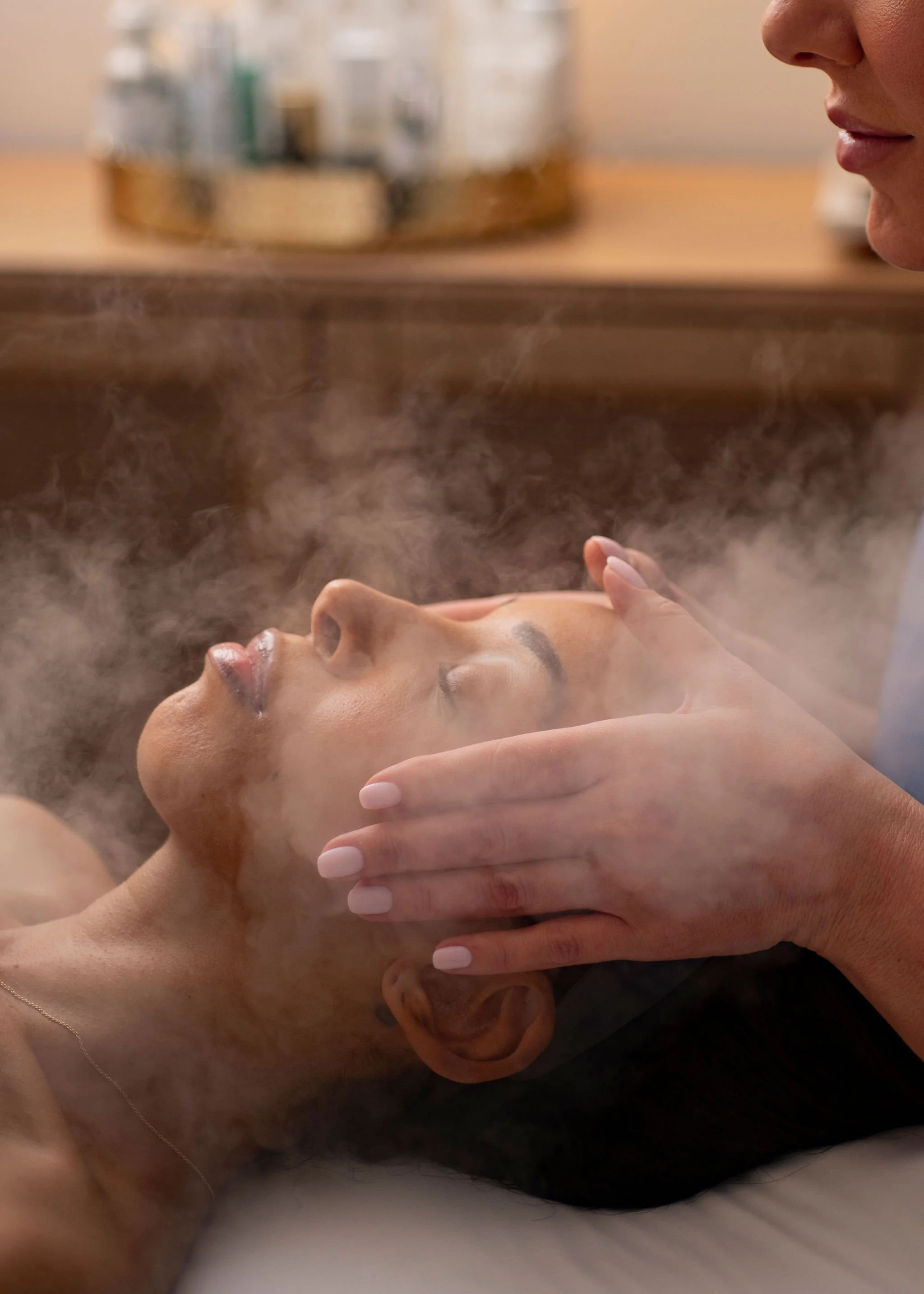 Two women in a medi spa setting. One woman is lying down with her eyes closed, and the other woman is gently holding her face and neck, performing a treatment. Steam or mist surrounds the scene.