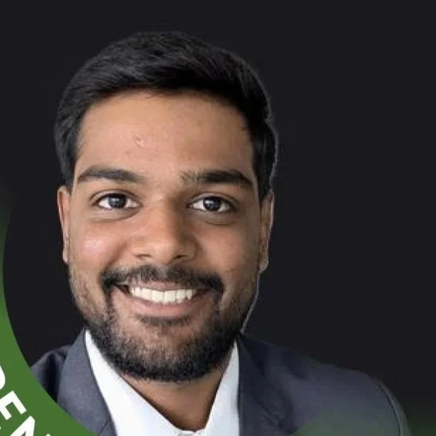 Portrait of a smiling man with dark hair and beard, dressed in a suit and white shirt, against a dark background.