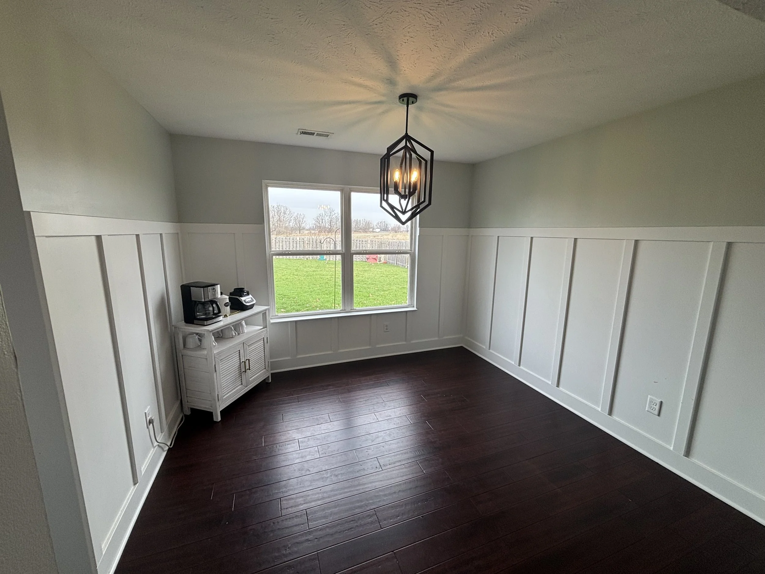 Empty dining room with dark wood flooring, white wainscoting walls, and a large window showing a backyard with grass and a wooden fence, hanging black geometric chandelier, and a small white cabinet with coffee maker and phone.