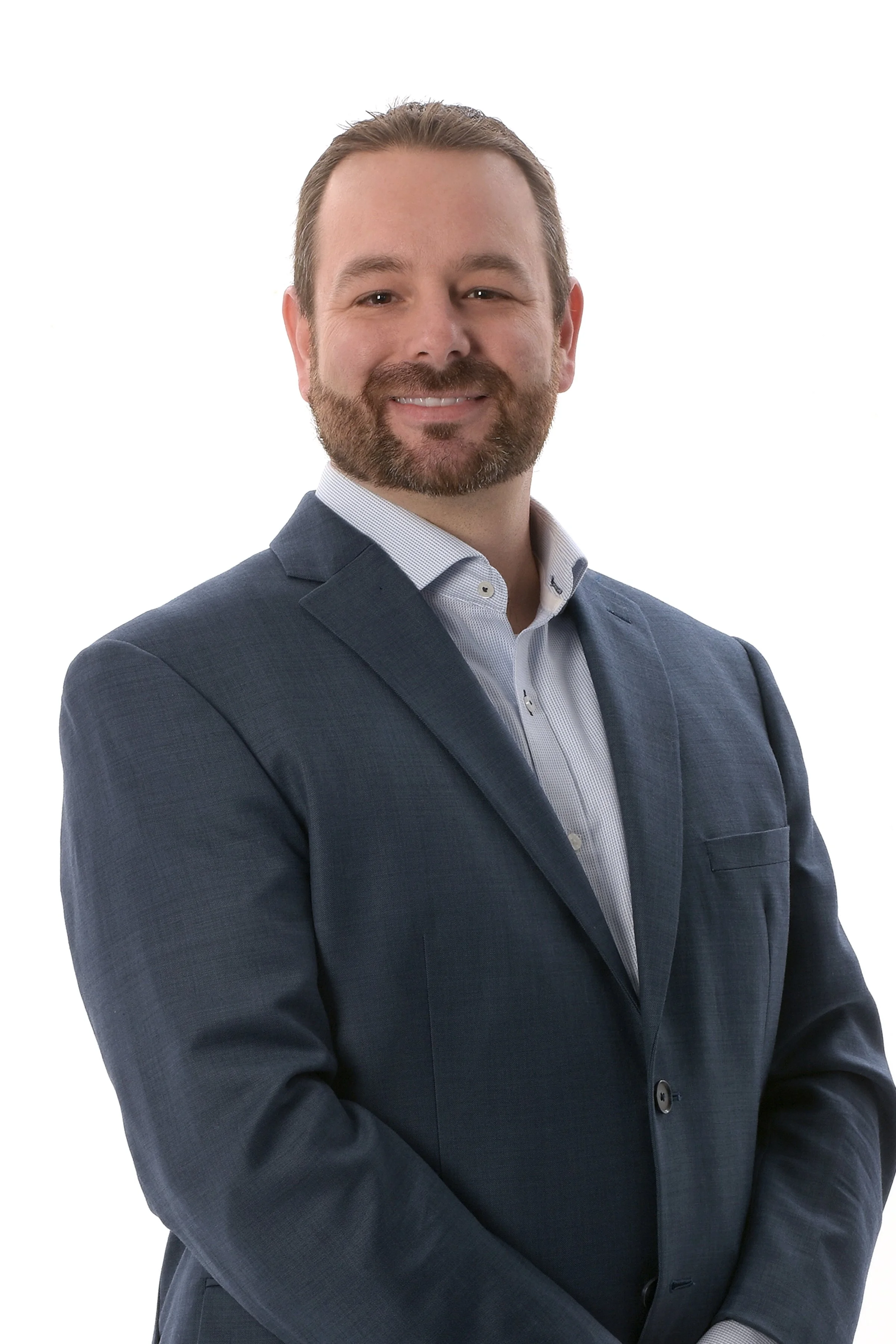 A man with a beard and brown hair wearing a navy blue suit and a white dress shirt, smiling with arms crossed against a white background.