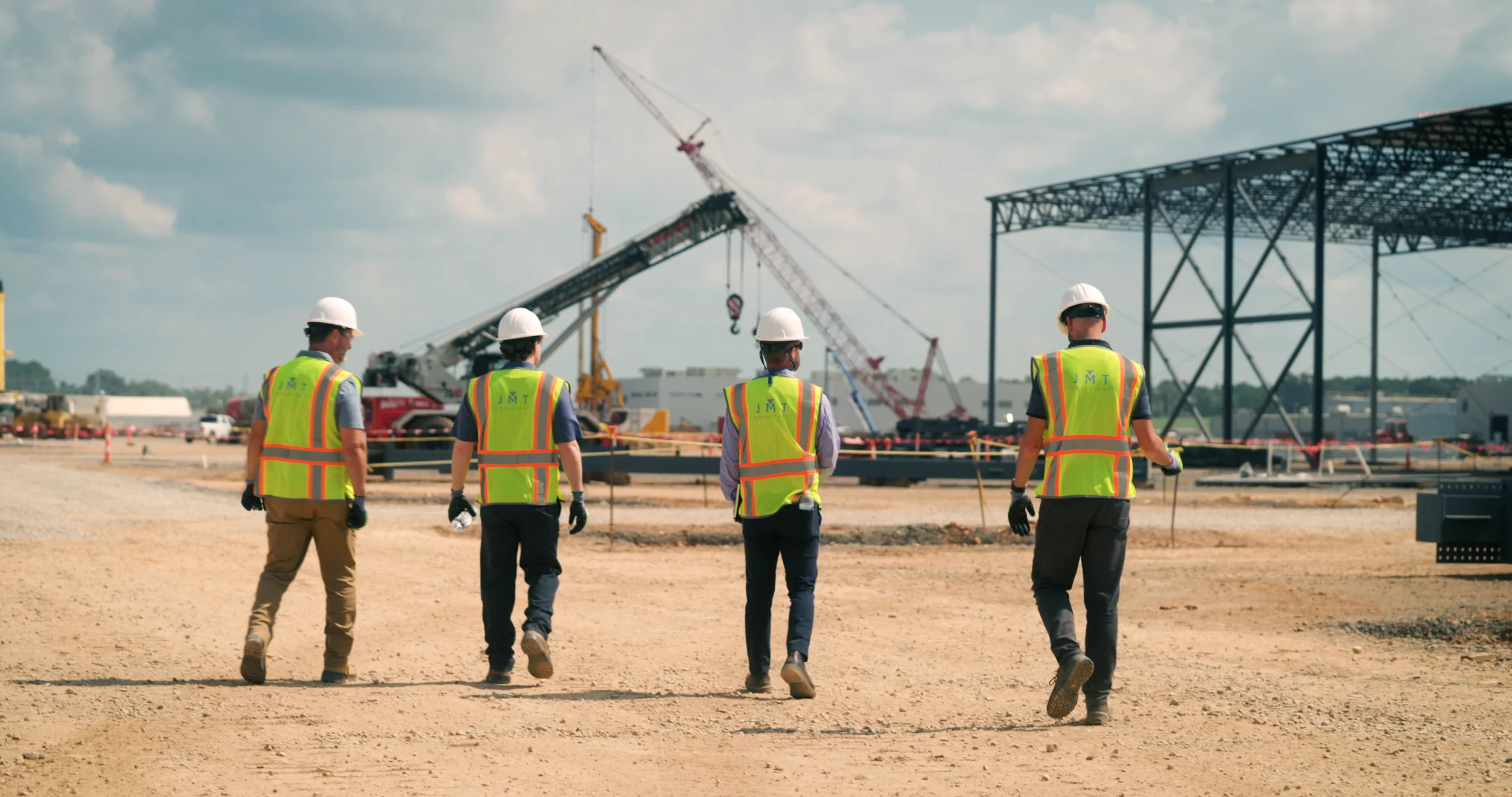 Four construction workers in safety vests and hard hats walking on a construction site, with cranes and metal structures in the background.