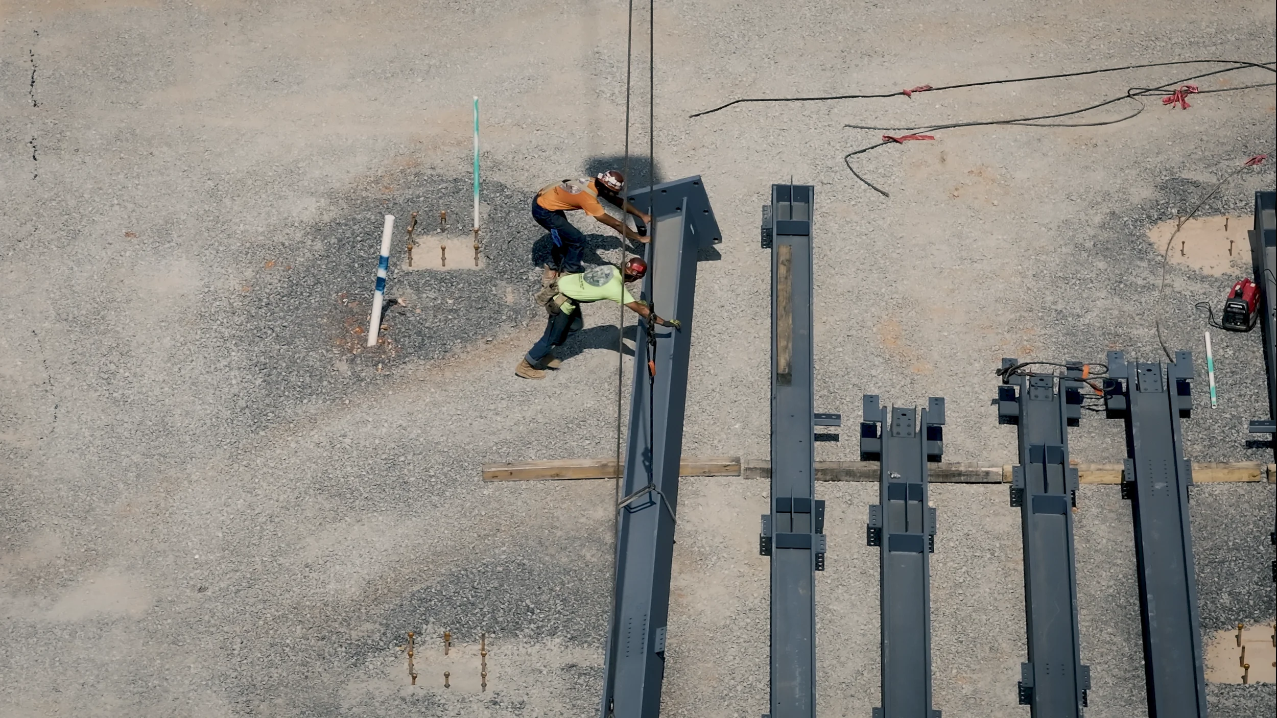 Two construction workers are assembling steel beams at a construction site on a gravel surface, with various equipment and wires nearby.