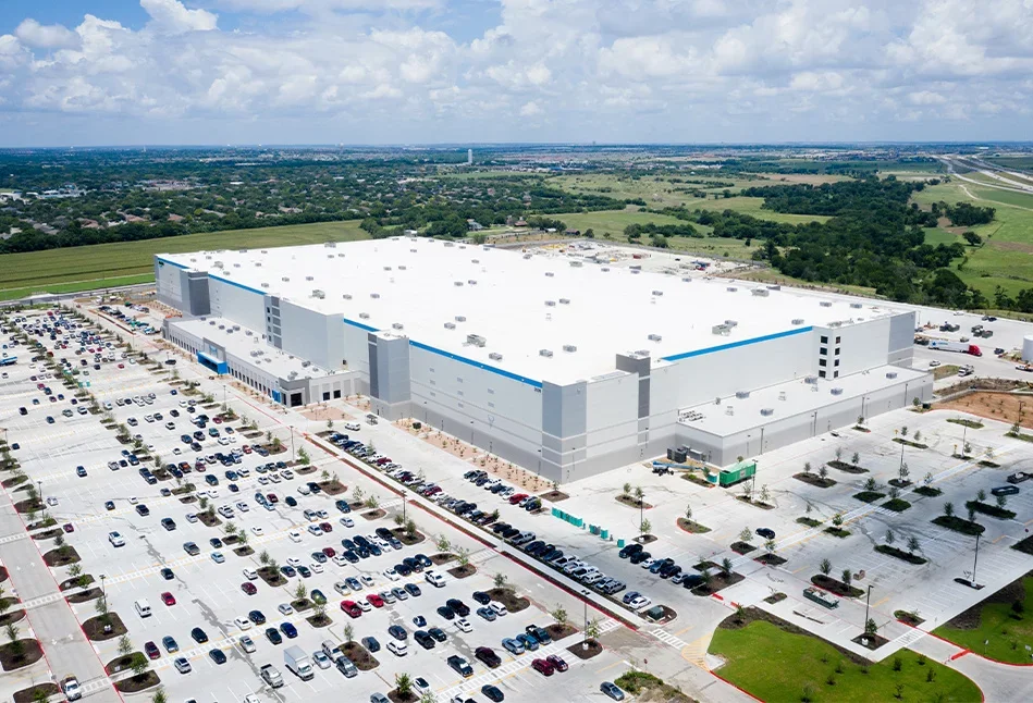 An aerial view of a large commercial warehouse with a surrounding parking lot filled with cars, set in a green, rural landscape under a partly cloudy sky.