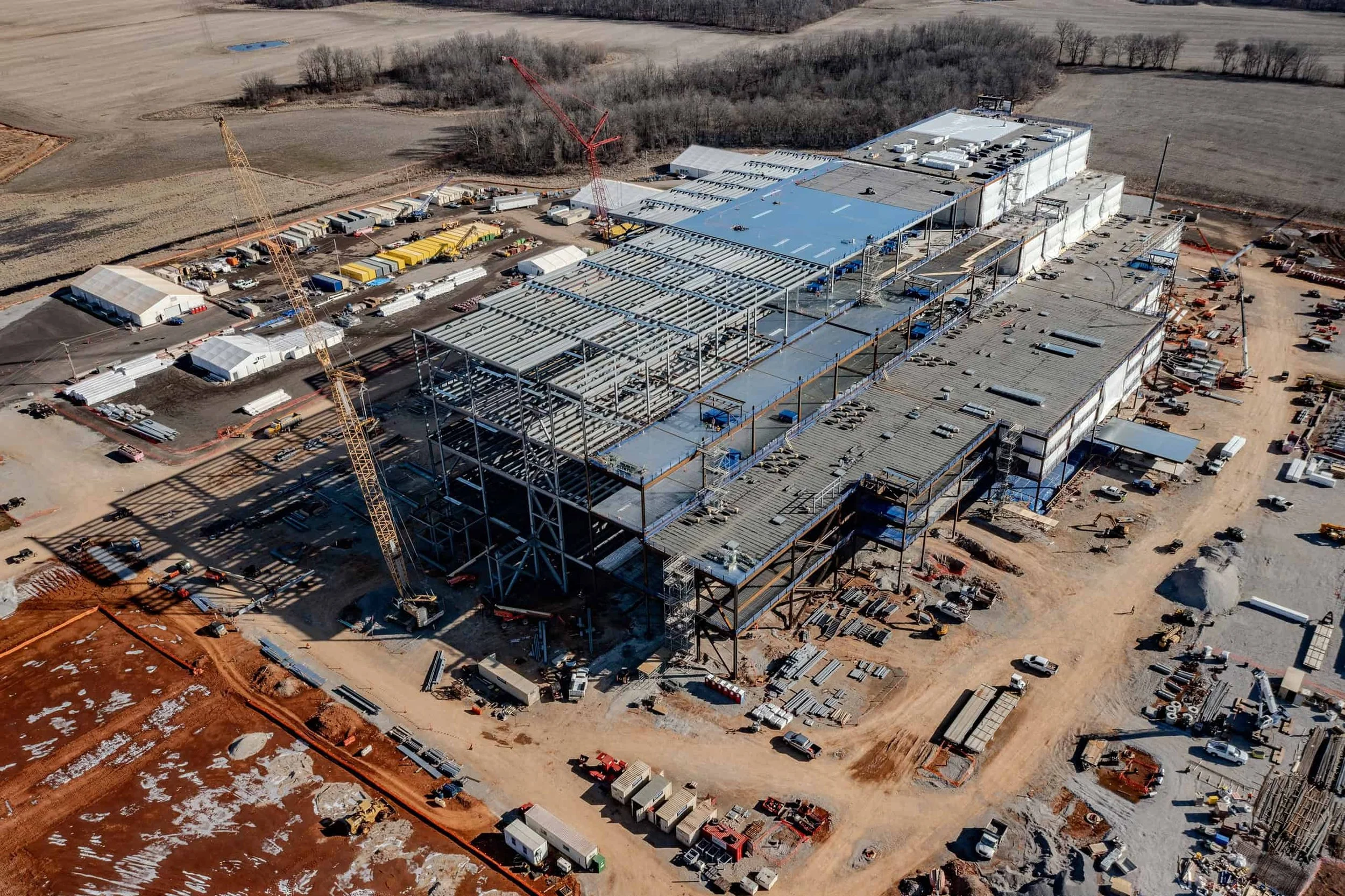 An aerial view of a large construction site with a partially built commercial building, cranes, construction vehicles, and materials scattered around, surrounded by open land and fields.