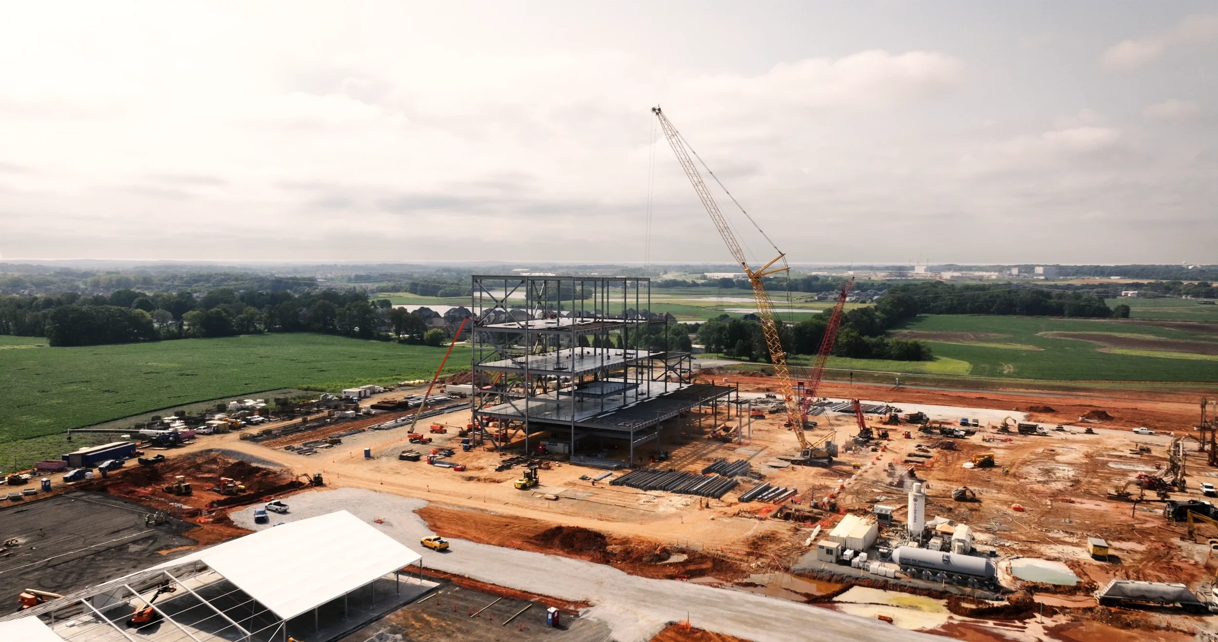 Construction site with a steel framework being built, cranes in use, and vehicles on dirt roads, surrounded by green fields and distant trees under a partly cloudy sky.