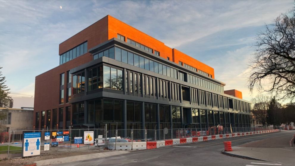 Modern multi-story building under construction, with orange and gray exterior, large glass windows, and construction barriers and signs in front.