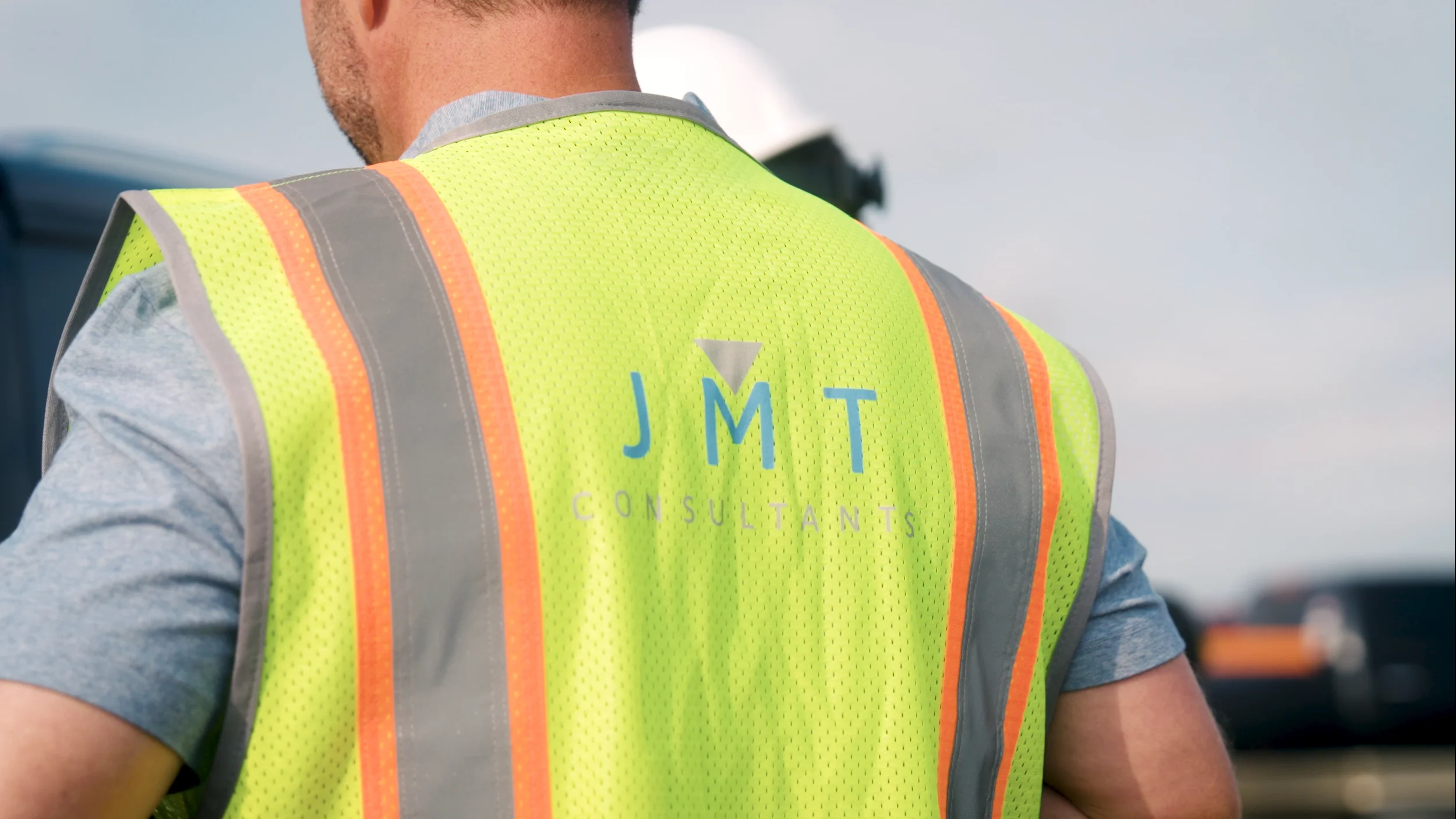 Close-up of a person wearing a yellow safety vest with orange and gray reflective stripes, with the letters 'JMT' and 'CONSULTANTS' printed on the back.