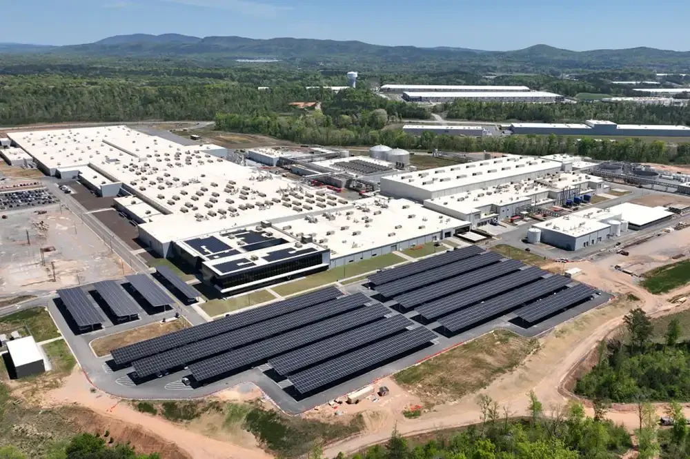 Aerial view of a large industrial building with solar panels in the foreground, surrounded by trees and mountains in the background.