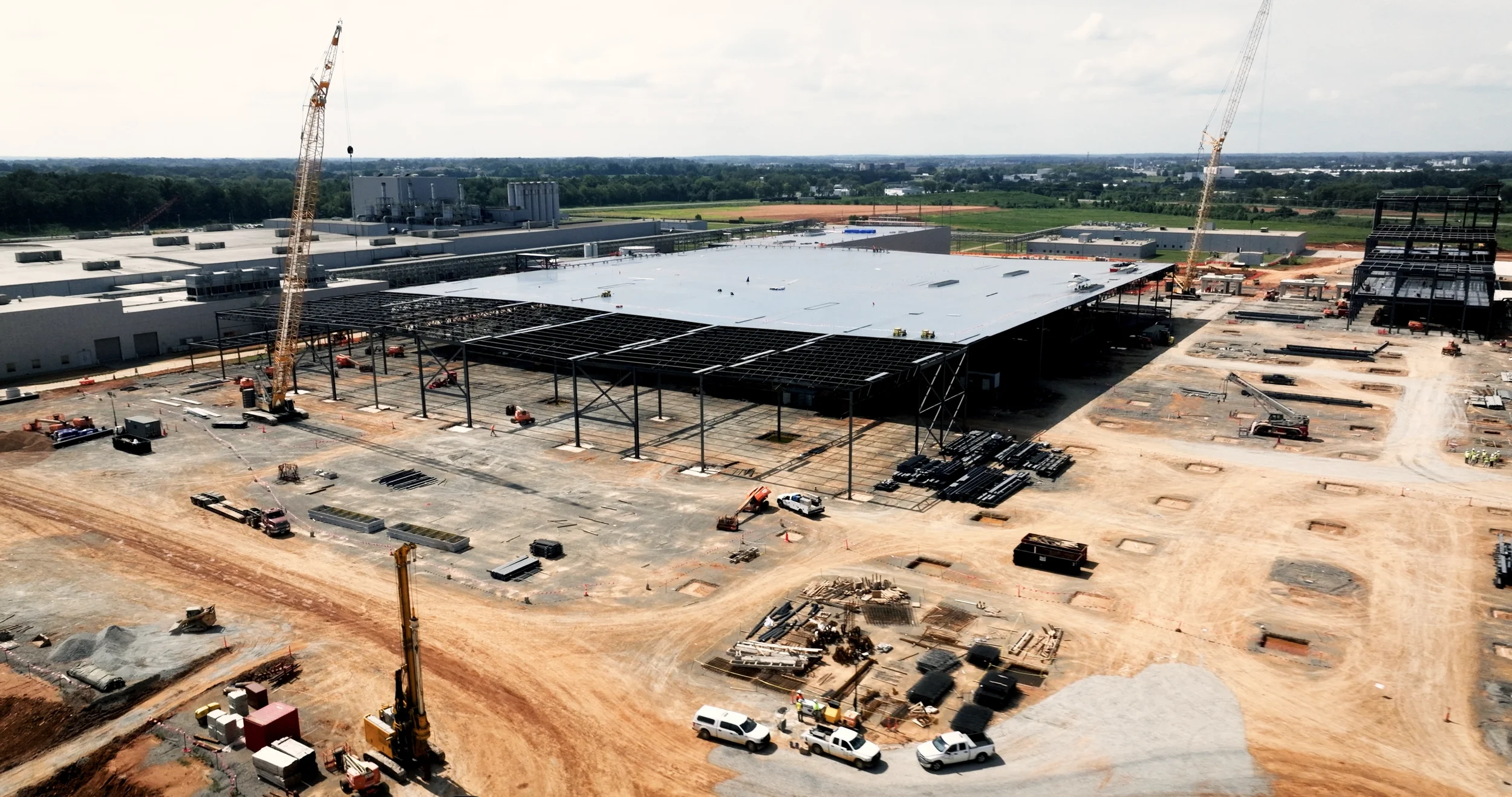 Construction site with cranes and steel framework for a large building, partially completed, surrounded by dirt and construction vehicles.