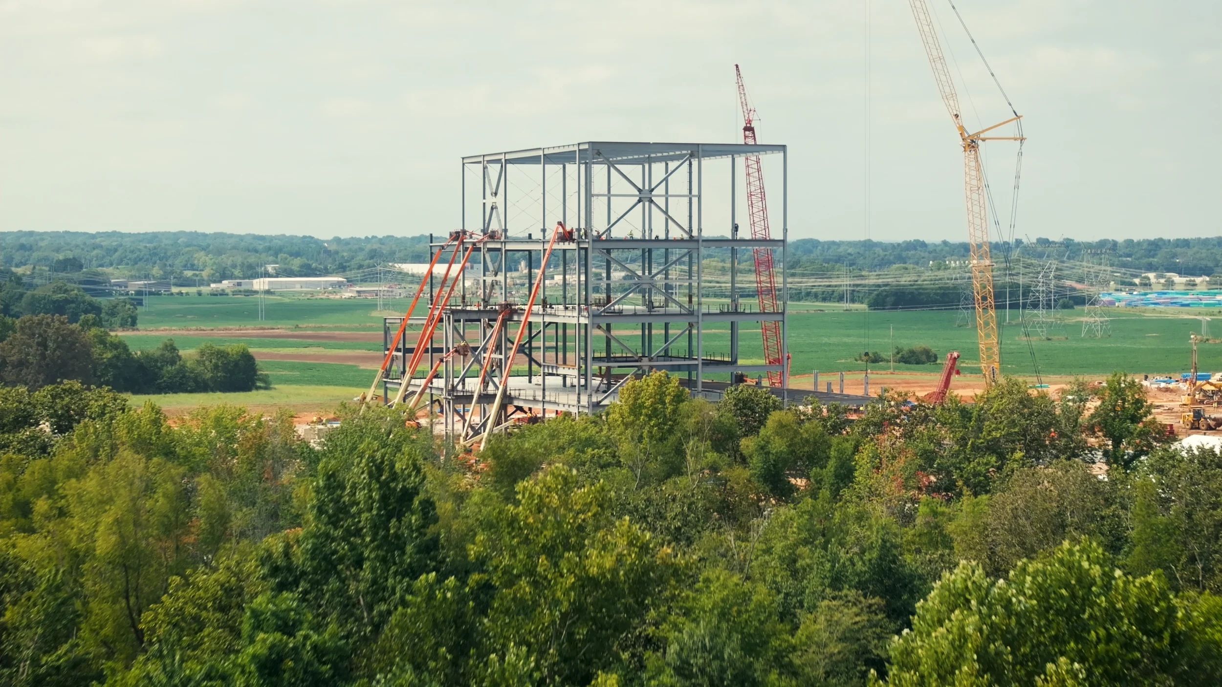 Construction site with a metal framework structure and cranes, surrounded by trees and green fields.