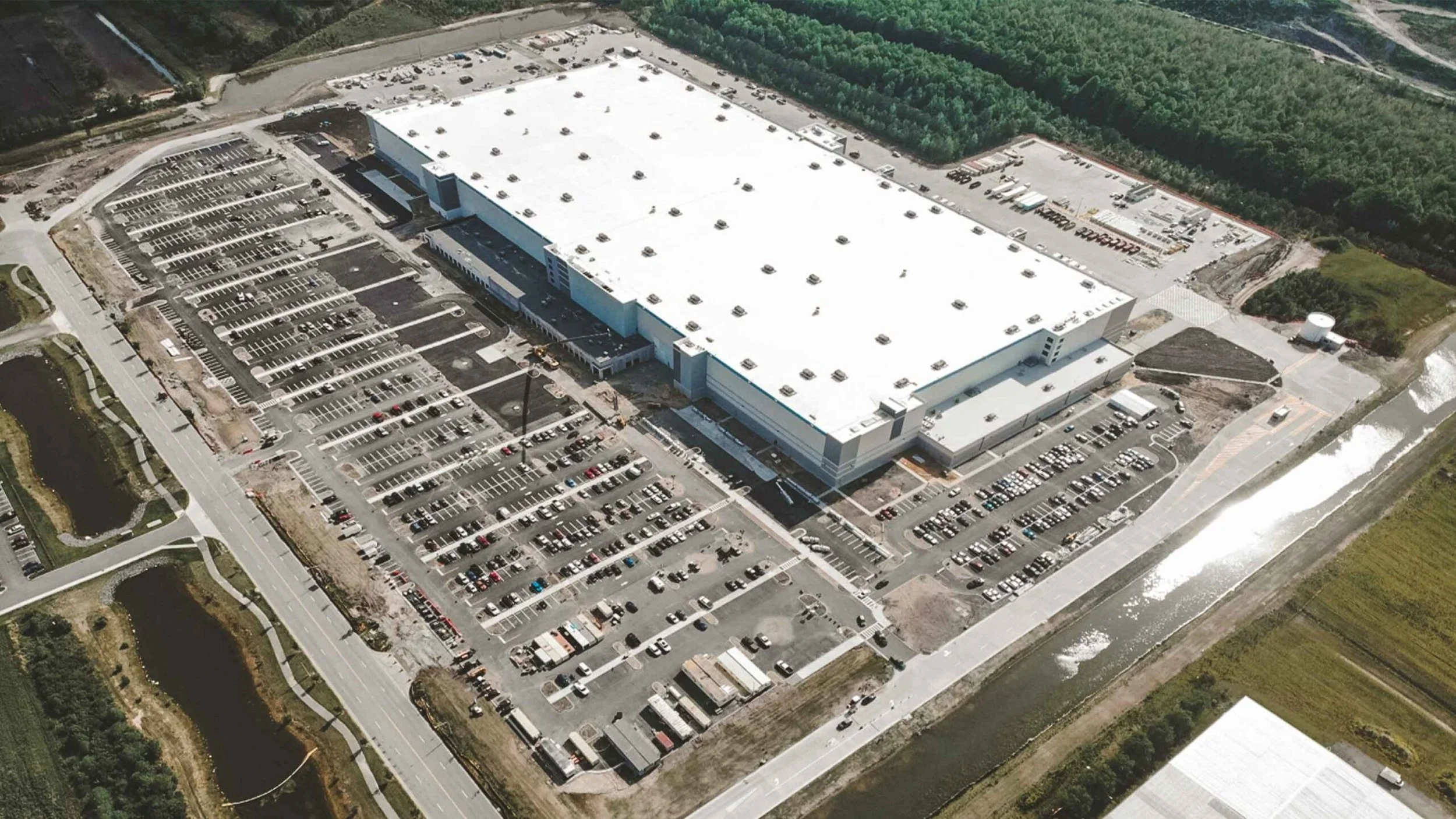 An aerial view of a large warehouse building surrounded by parking lots filled with cars and trucks, with a green forest in the background.