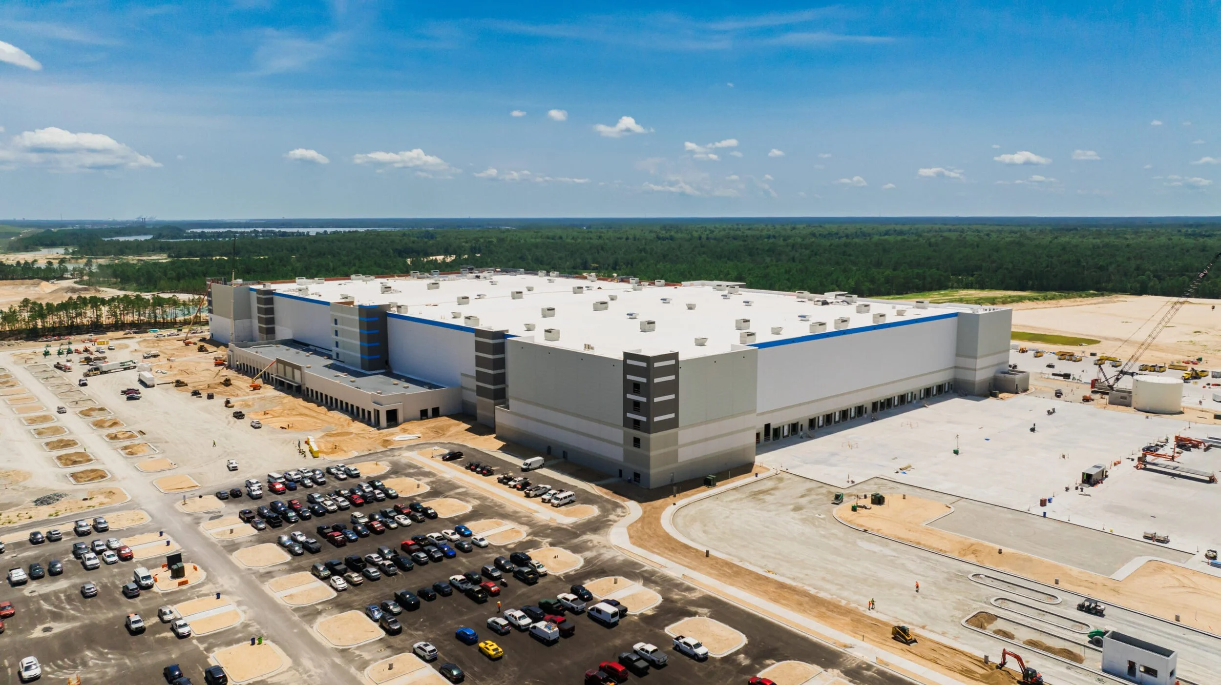An aerial view of a large upcoming retail or warehouse building with many parking spaces and construction activity around it, set in a green, rural landscape under a bright blue sky with scattered clouds.