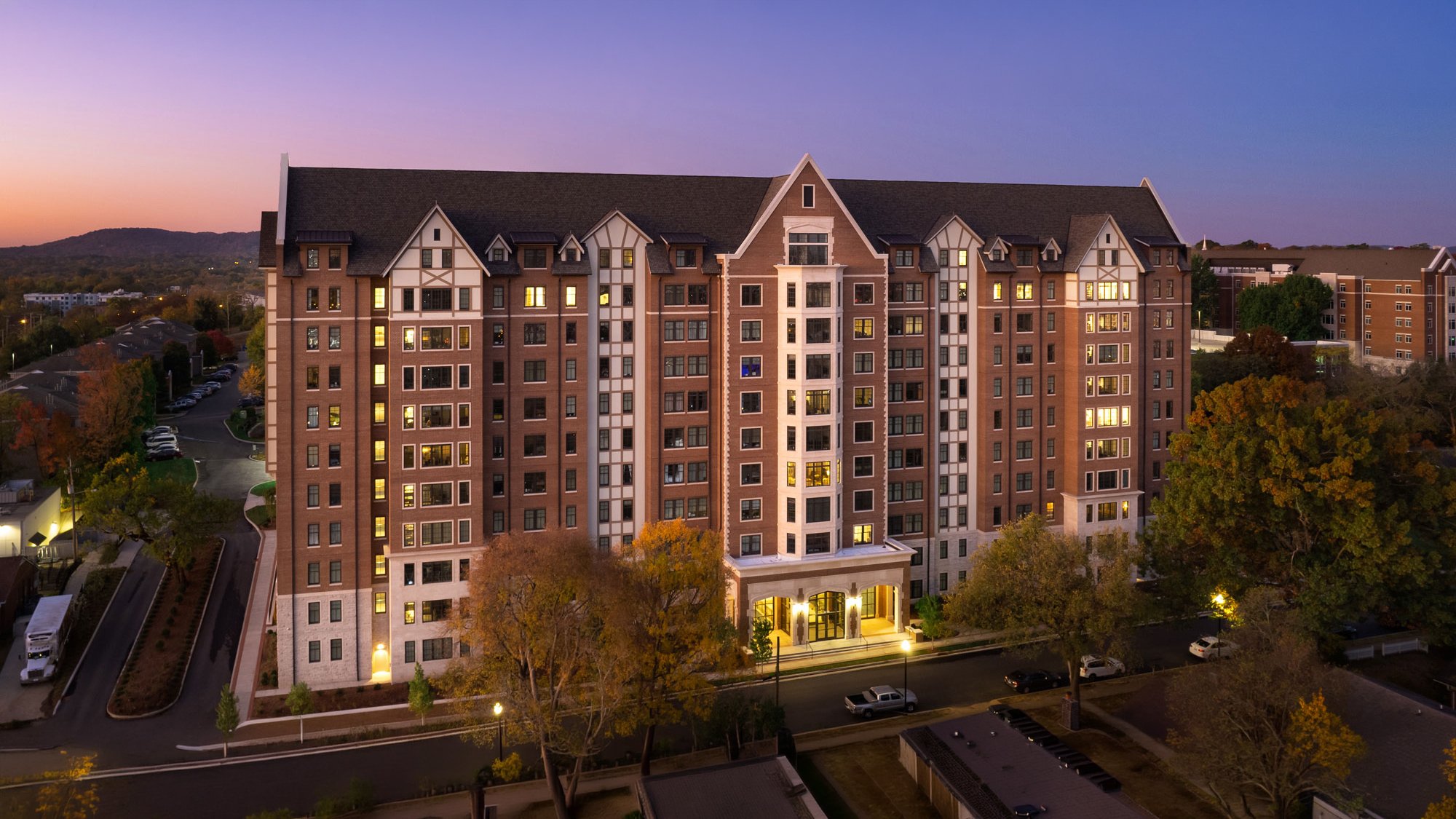 A large multi-story apartment building at dusk with lights on in many windows. The building has a brick exterior with white accents and a steep, dark roof. Surrounding trees and a parking lot are visible in the foreground, with a hilly landscape in the background.