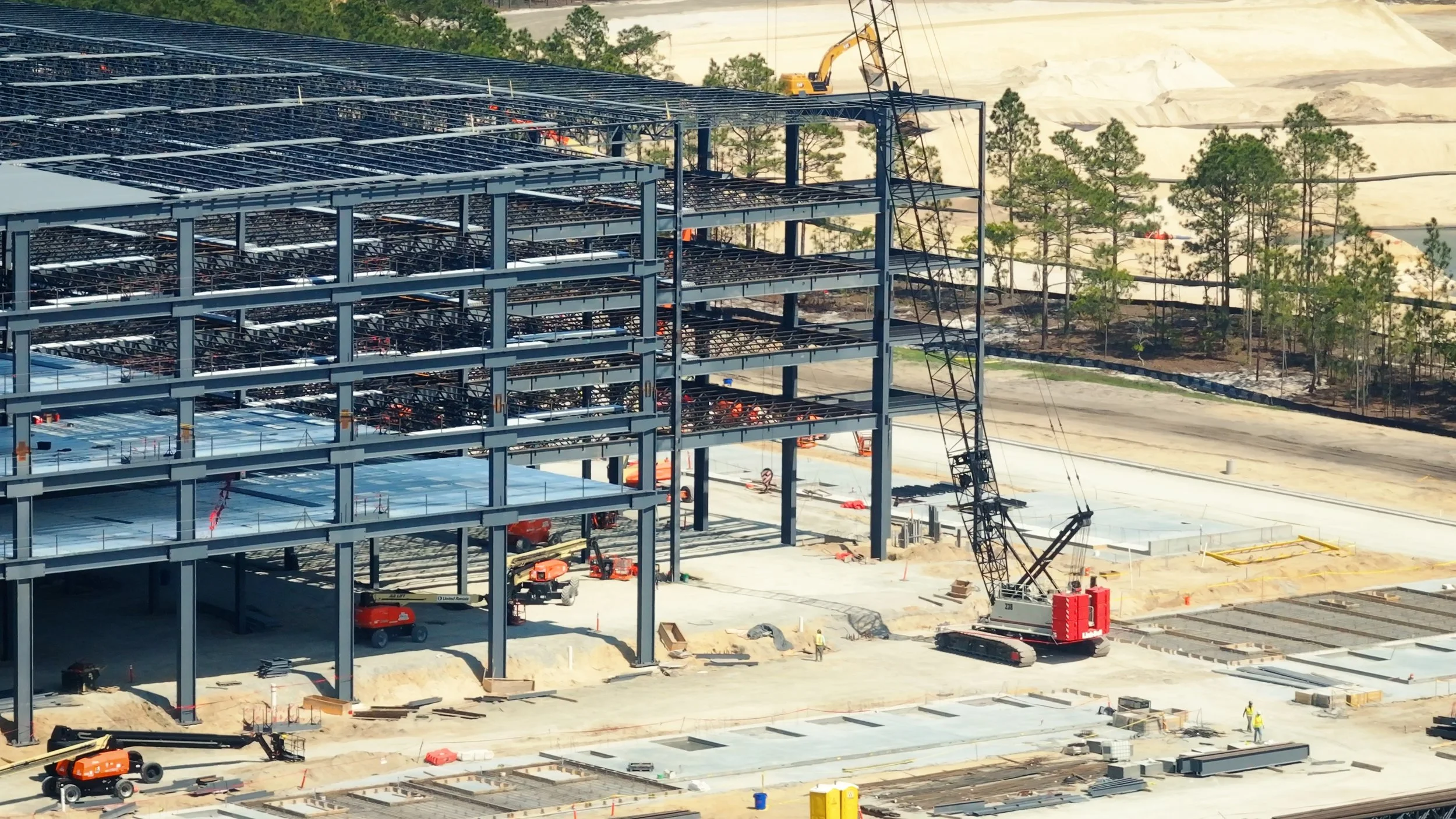 Construction site with a multi-story steel building frame, cranes, construction vehicles, and workers, surrounded by trees and open land.