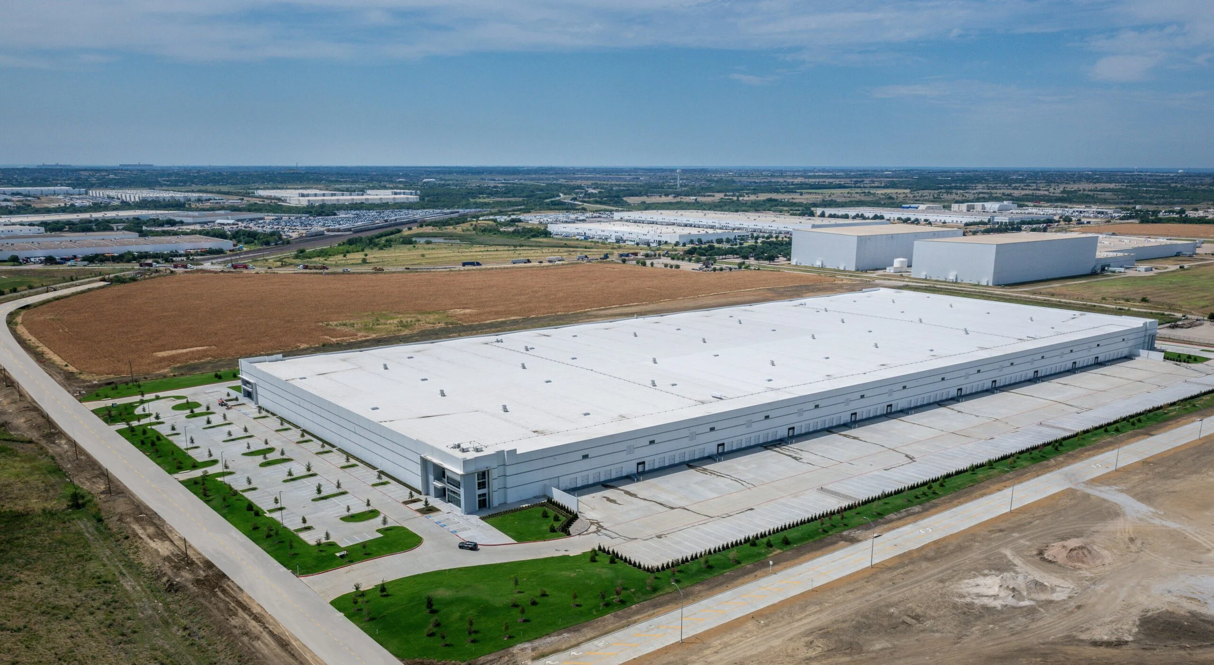 Aerial view of a large warehouse complex with a white rooftop, landscaped parking area, and surrounding open land, with additional industrial buildings and parking lots in the distance.