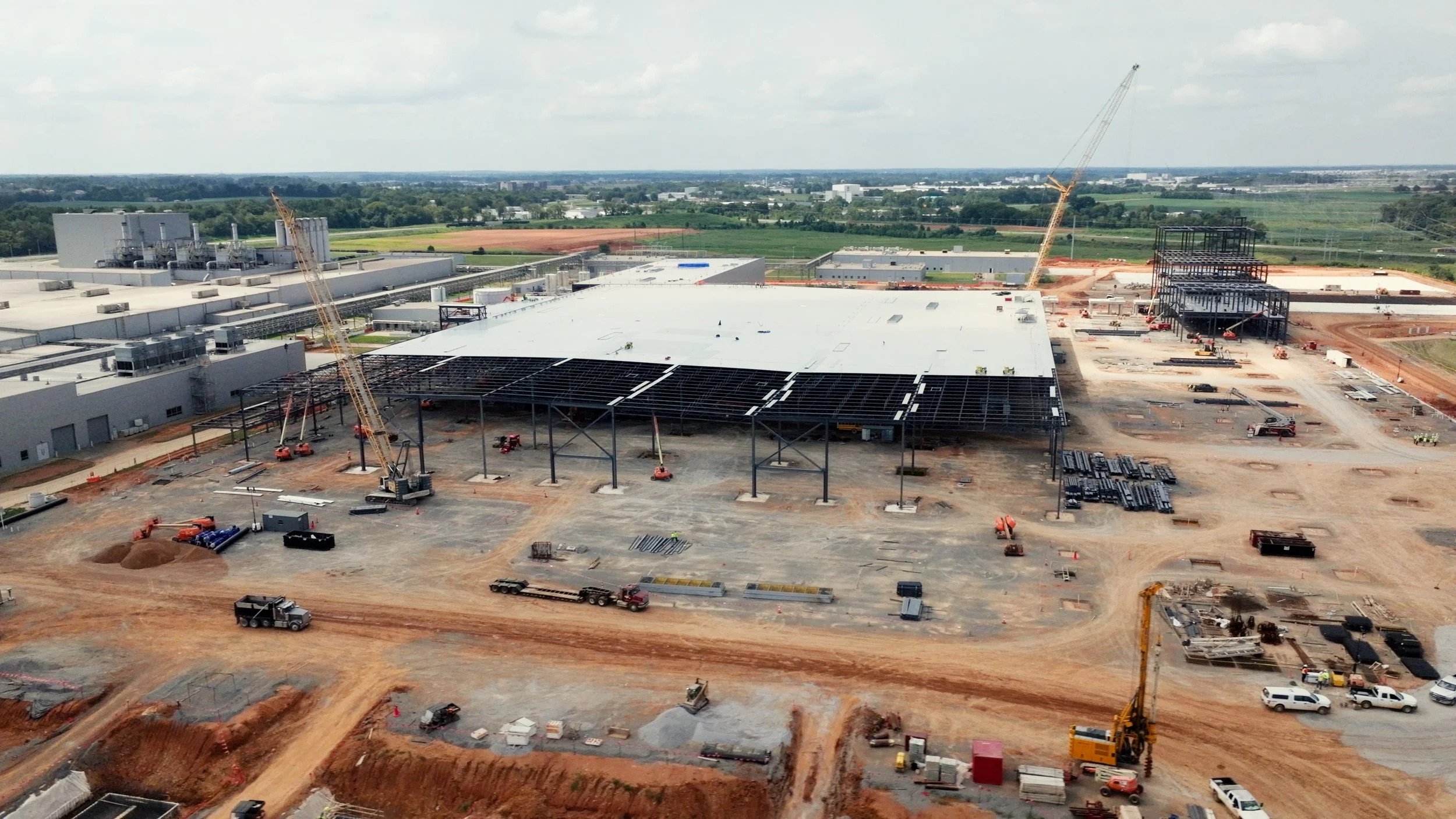 Under construction industrial building with cranes, construction vehicles, and steel framework, surrounded by dirt roads and open fields.