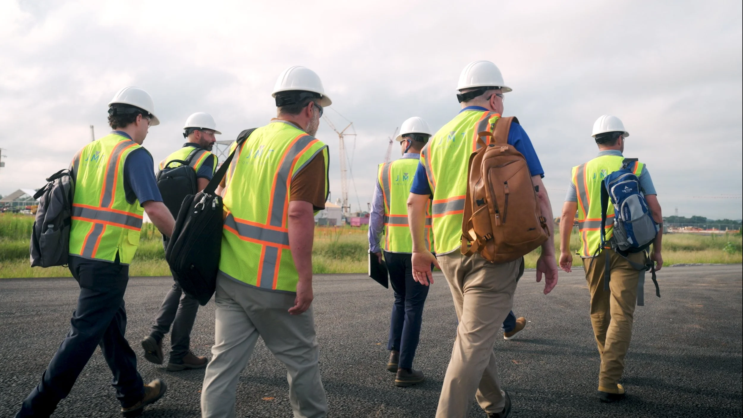 Group of construction or engineering professionals walking on a project site, wearing safety vests and hard hats, with construction cranes and equipment in the background.