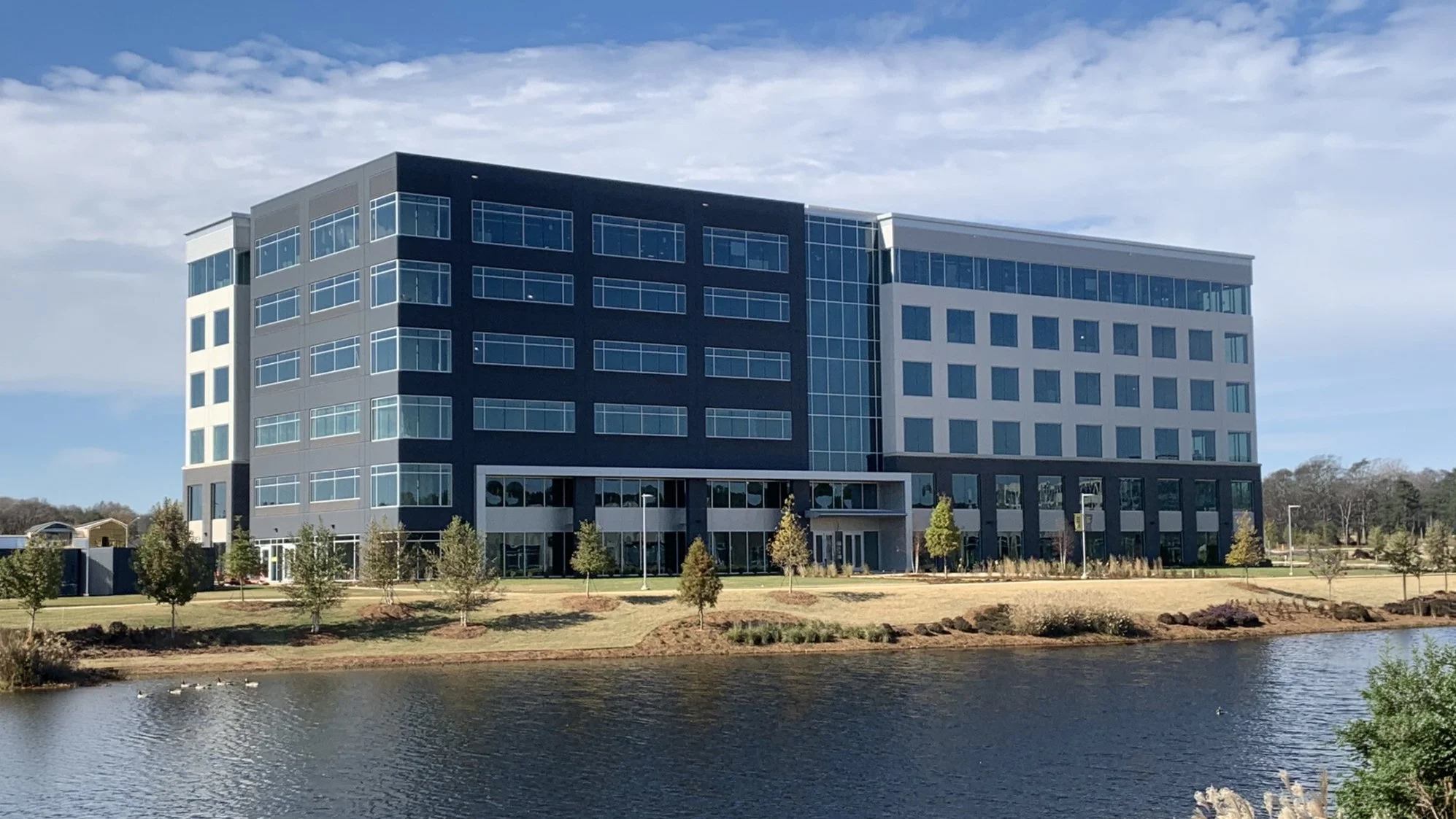 A modern multi-story office building with glass windows, situated near a water body with trees and shrubs in the landscape.