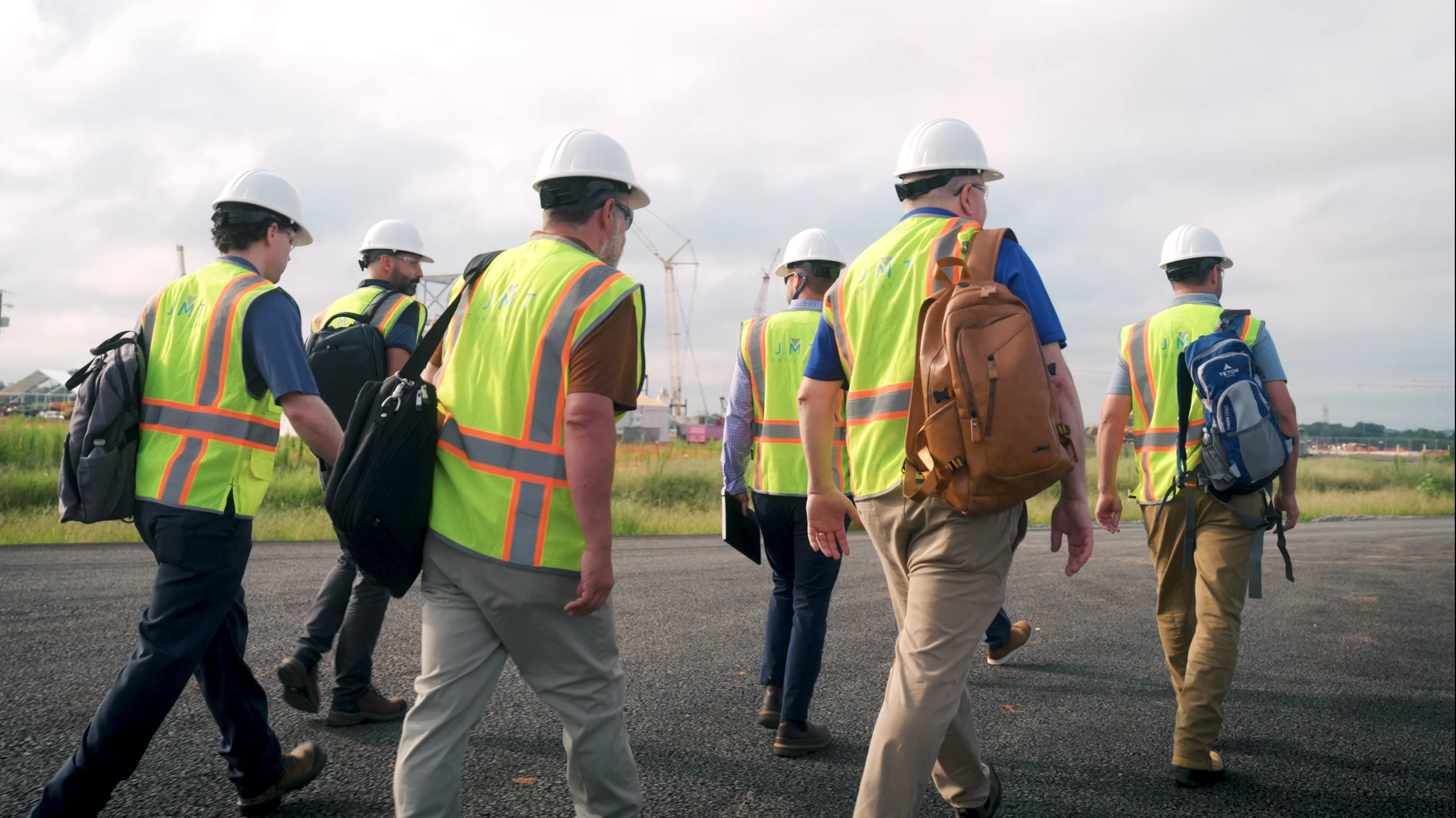 A group of people wearing safety vests and white hard hats walking on a construction site with cranes and buildings in the background.