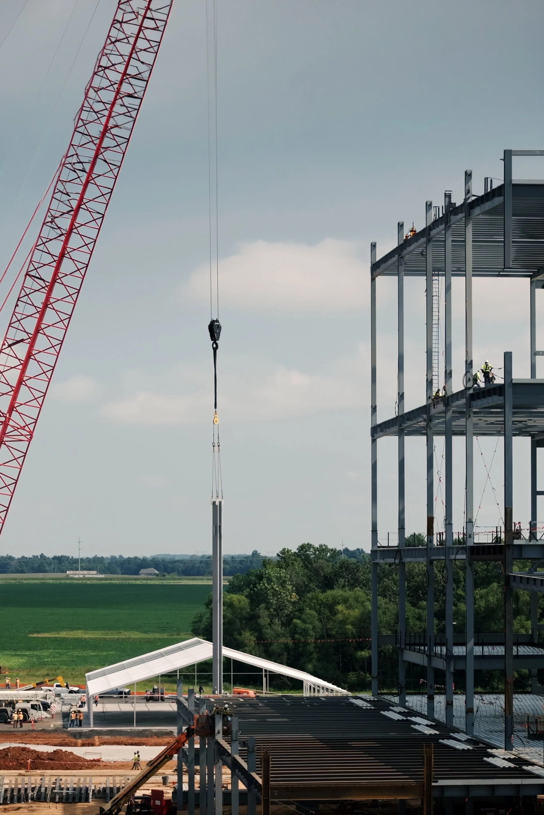Construction workers building a multi-story building with a steel frame, construction crane in the background, and a rural landscape with trees and fields in the distance.