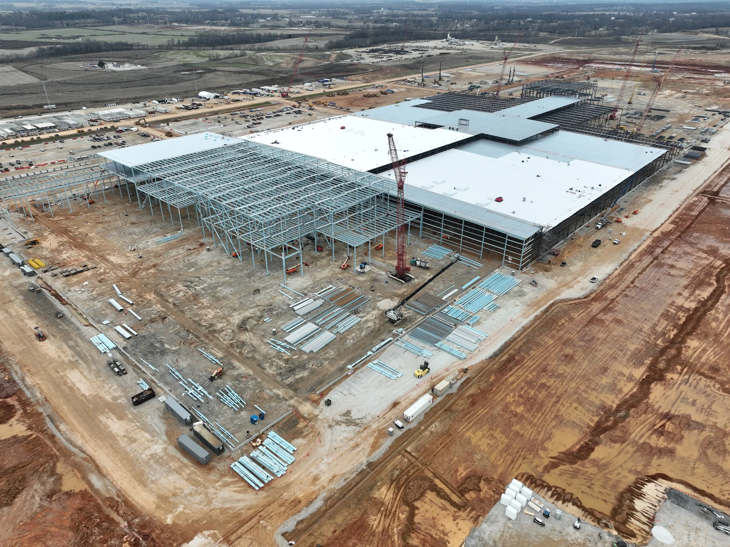 Aerial view of a large construction site with steel framework and partially built structures, cranes, construction materials, and vehicles on dirt roads.