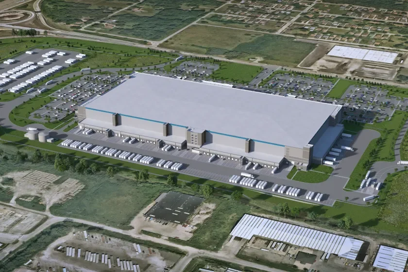 A large industrial warehouse with multiple loading docks, trucks parked outside, and surrounding parking lots and greenery.