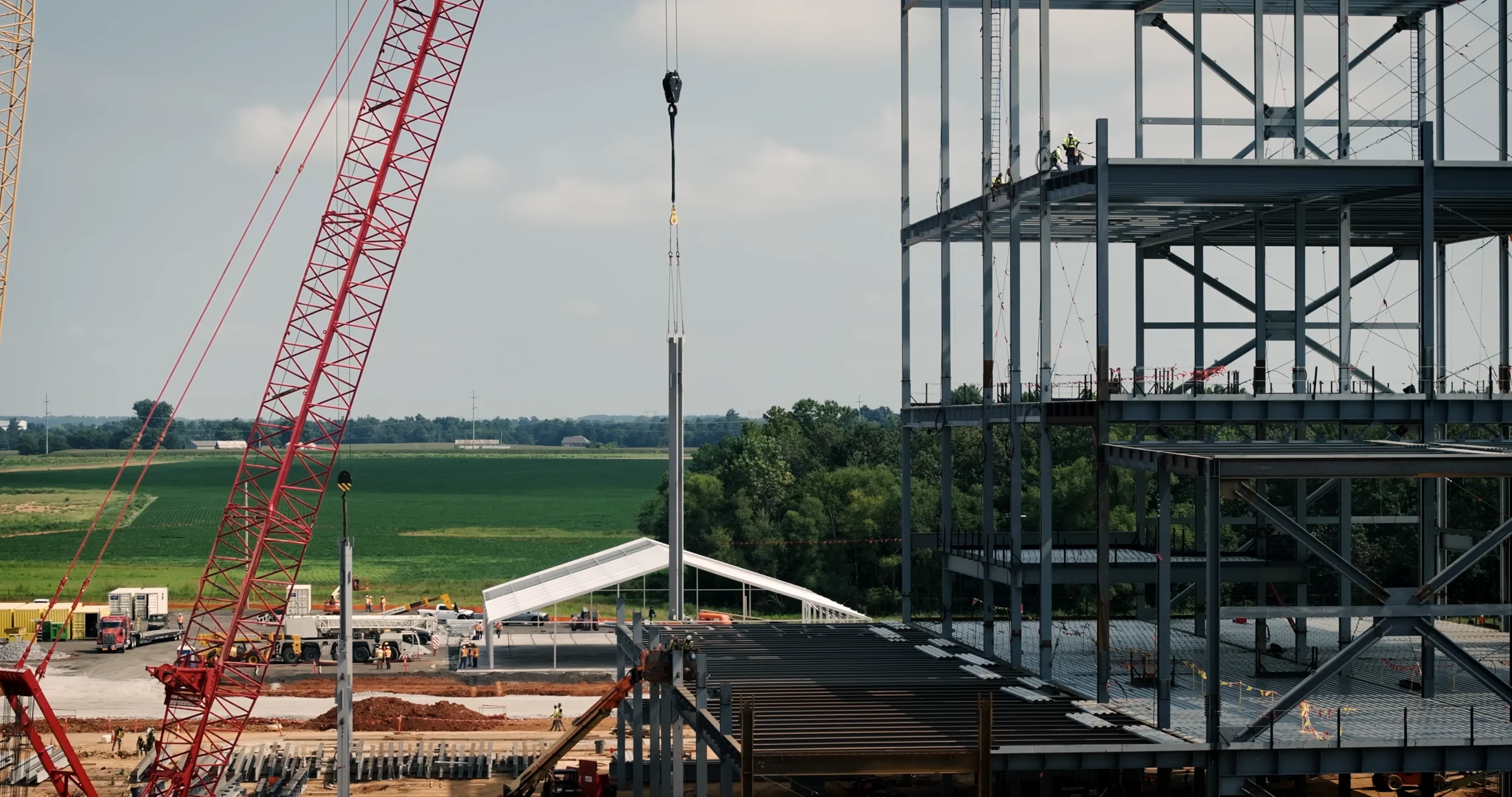 Construction site with a large red crane, steel framework of a building, construction workers, and machinery, with a background of green fields and a partly cloudy sky.