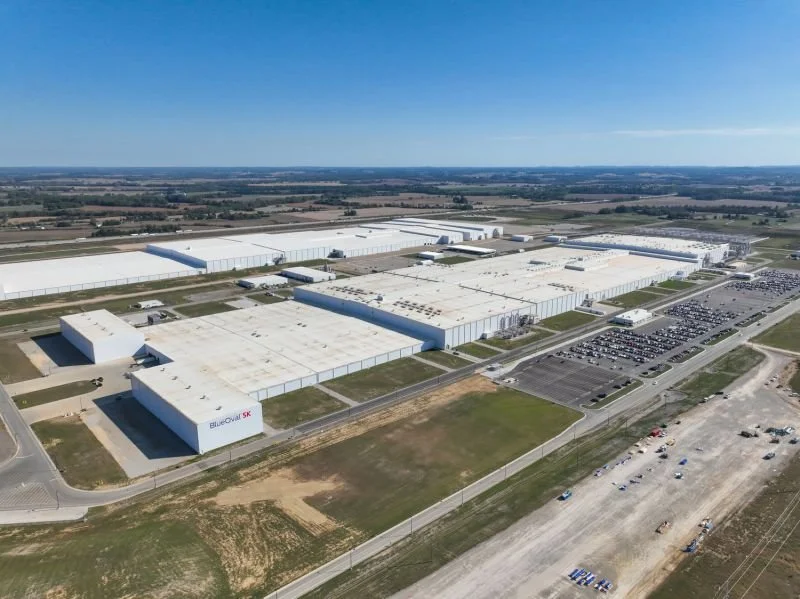 An aerial view of an industrial complex with multiple large white warehouse buildings, a parking lot filled with cars, and surrounding open fields under a clear blue sky.