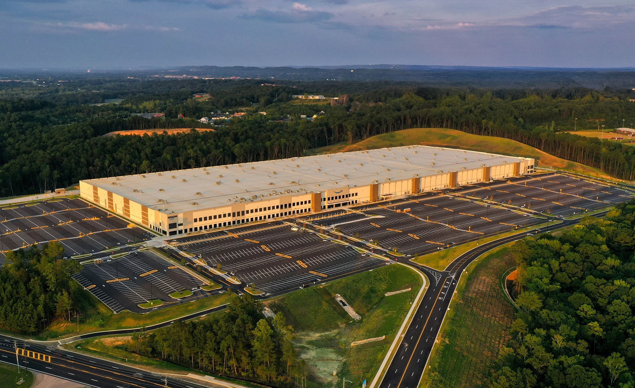 An aerial view of a large Amazon distribution warehouse surrounded by an extensive parking lot with multiple empty parking spots, green trees, and a hilly landscape in the background.