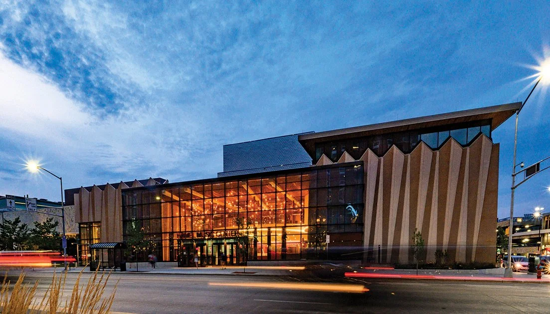 A modern building with a large glass front and illuminated interior, likely a museum, with a parking garage on the right, during evening with a partly cloudy sky.