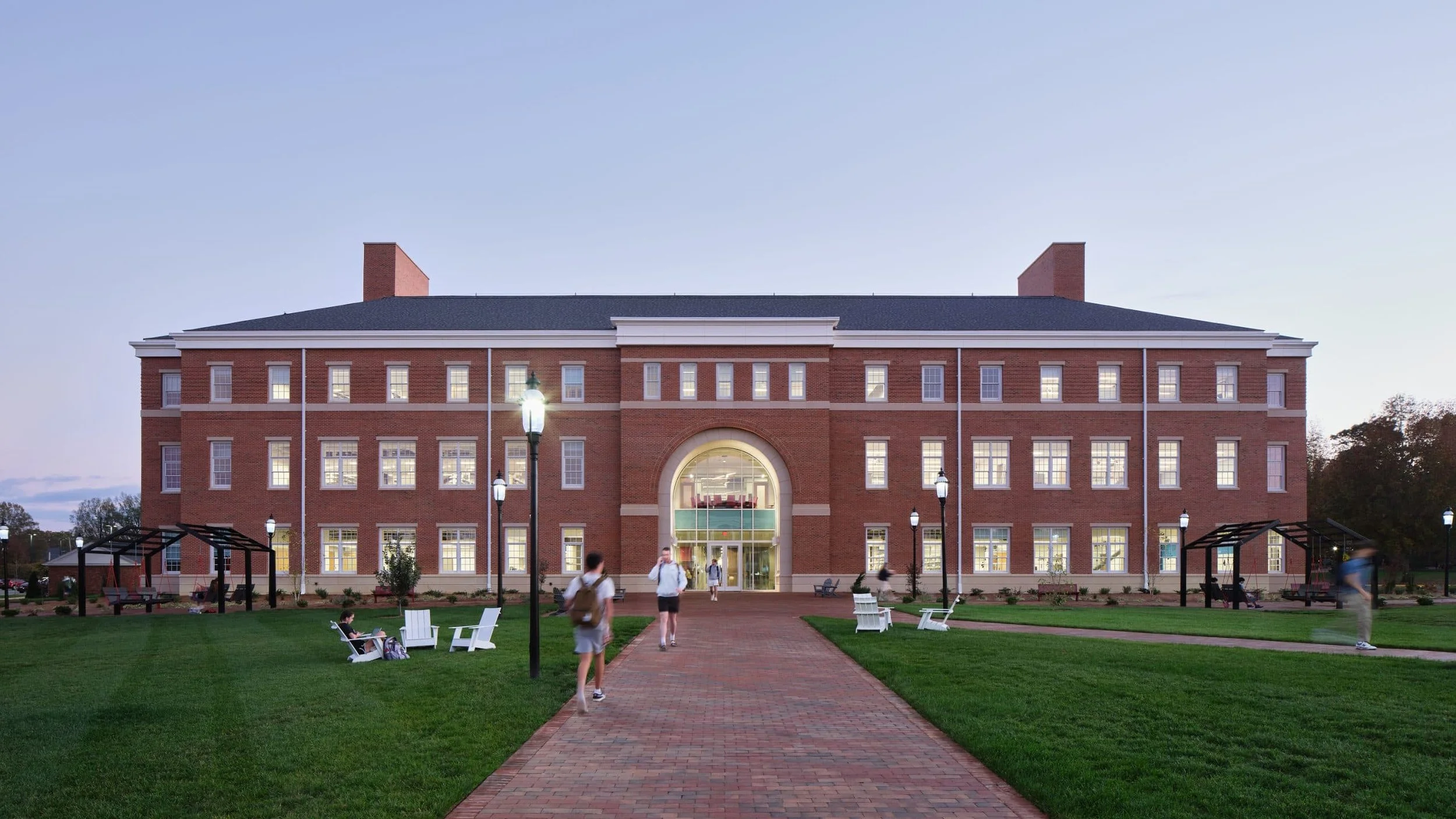A large brick building with multiple windows, a central archway entrance, and a green lawn with benches and sprouting trees in the front. Several people are walking or sitting around the area, and there are street lamps along the brick walkway leading to the entrance. The sky is clear with a hint of sunset light.