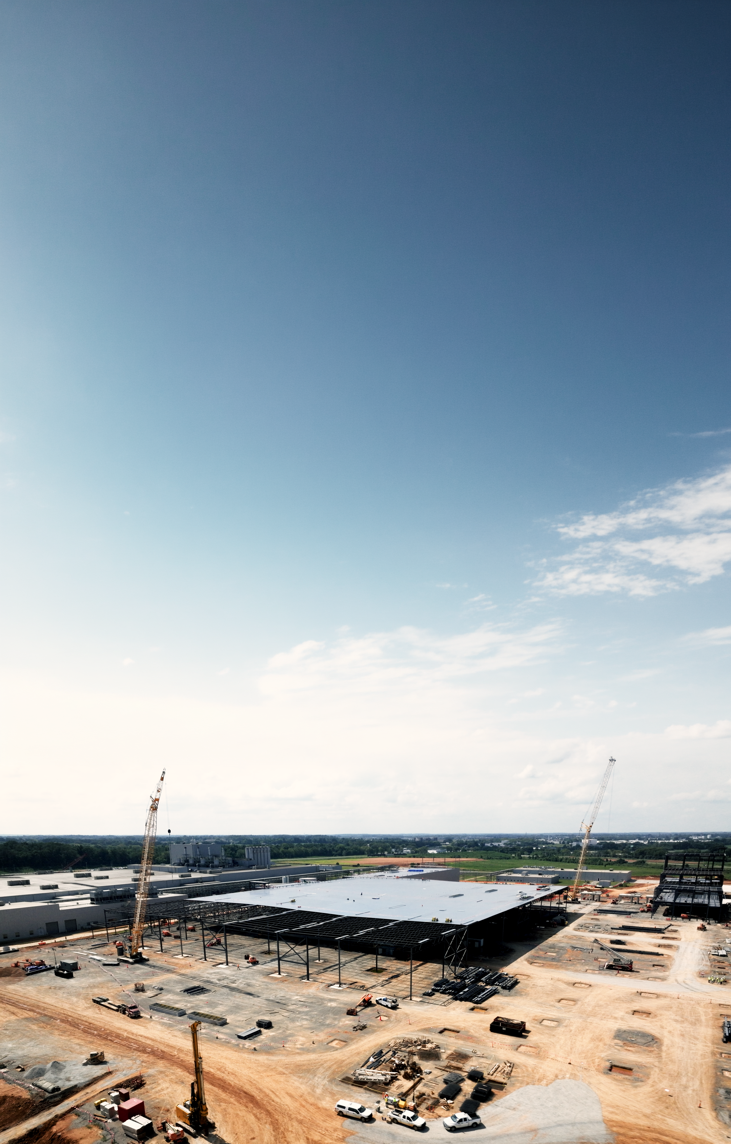 Construction site with cranes, partially built structure, building materials, and vehicles, under a blue sky with scattered clouds.
