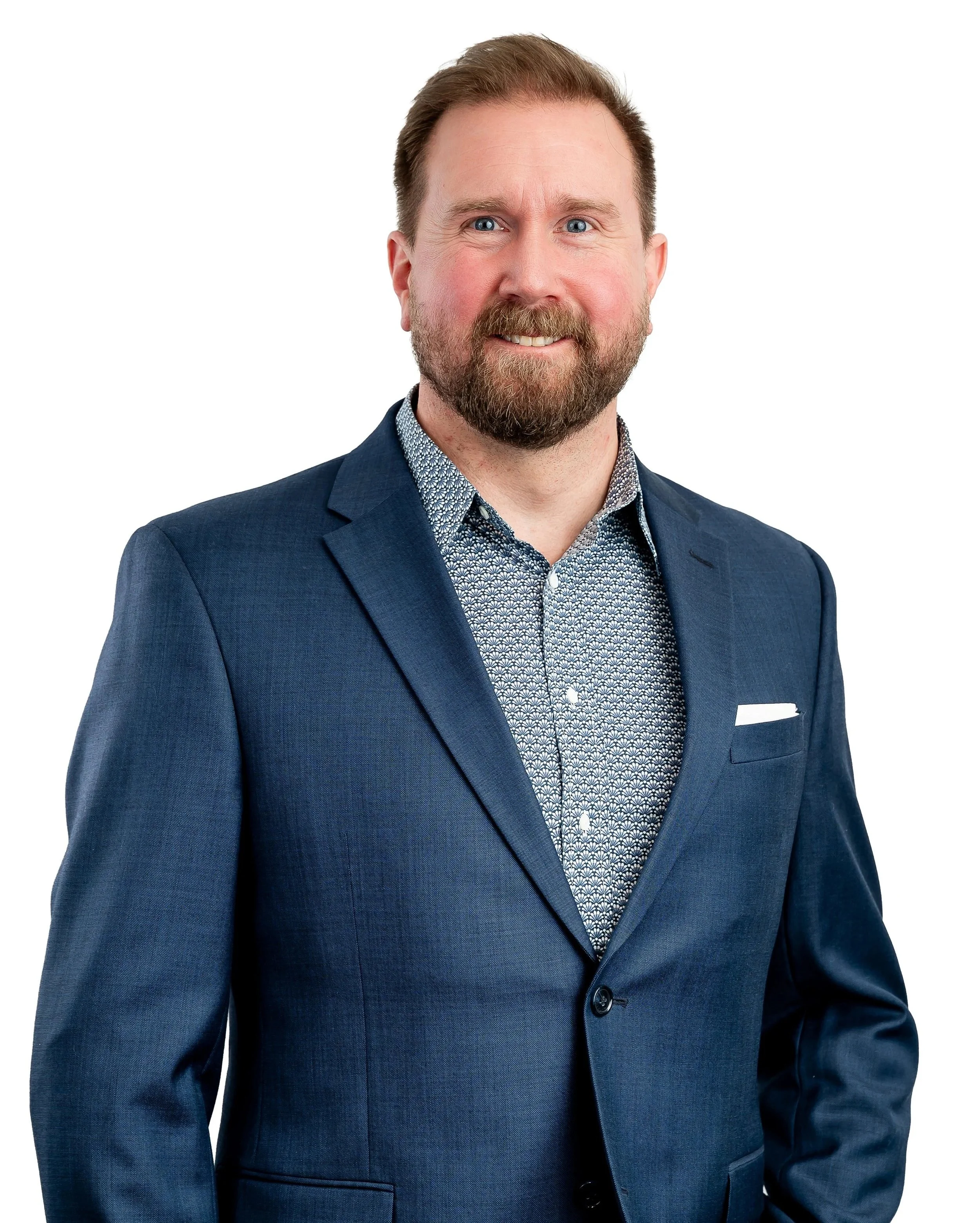 A man with light skin, blue eyes, reddish-brown hair, and a beard wearing a navy blue suit jacket over a patterned dress shirt against a white background.