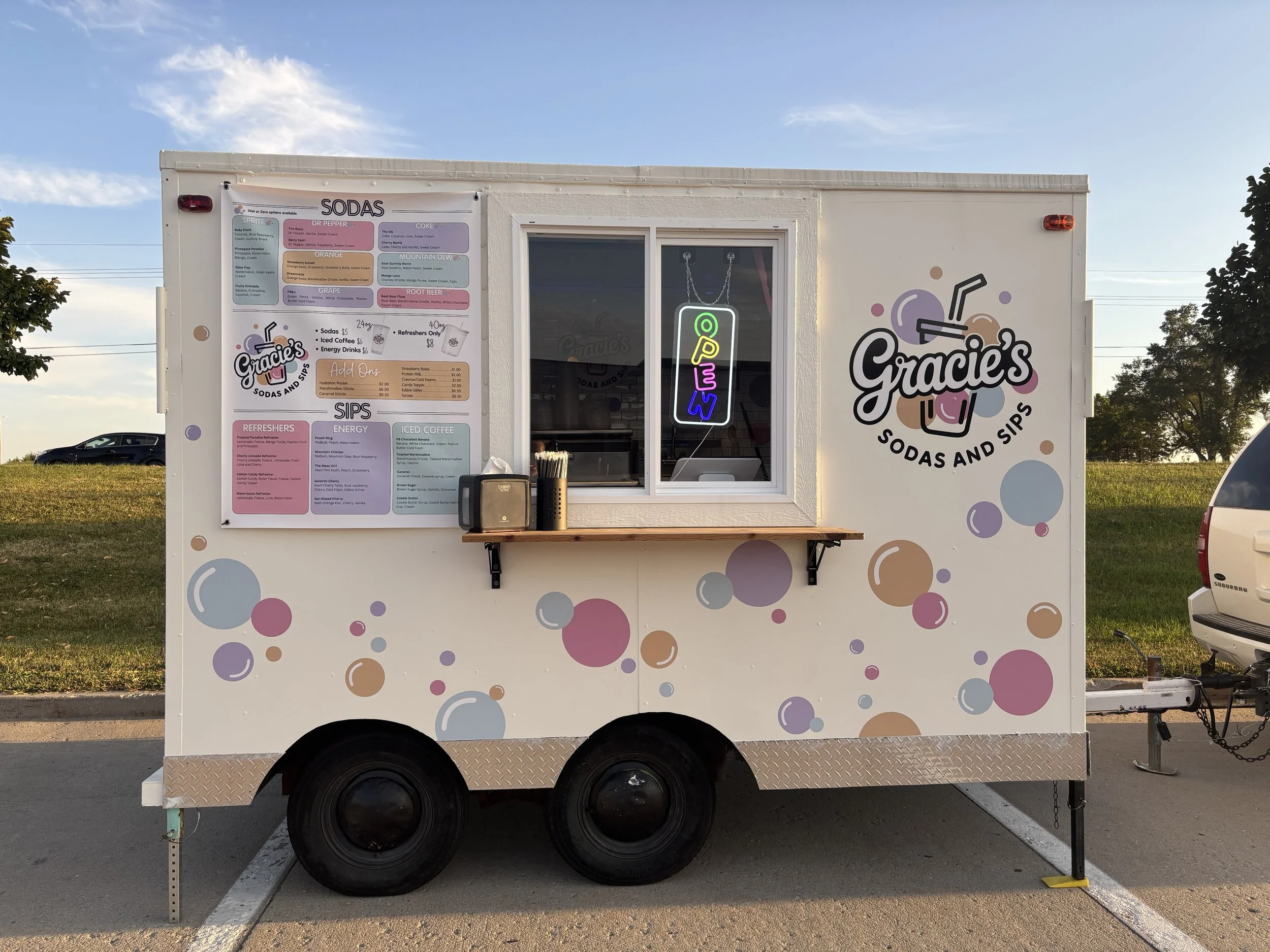 A white food trailer with colorful bubbles and a logo that says "Gracie's Sodas and Sips" parked in a lot. The trailer has a menu board, a window with a neon "OPEN" sign, and a small shelf with supplies.