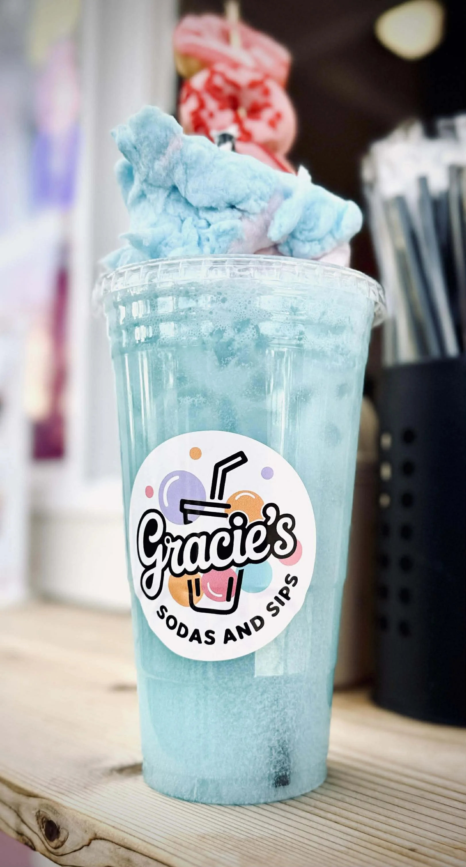 A blue cotton candy topped soda drink in a clear cup with a 'Gracie's Sodas and Sips' logo, placed on a wooden surface.