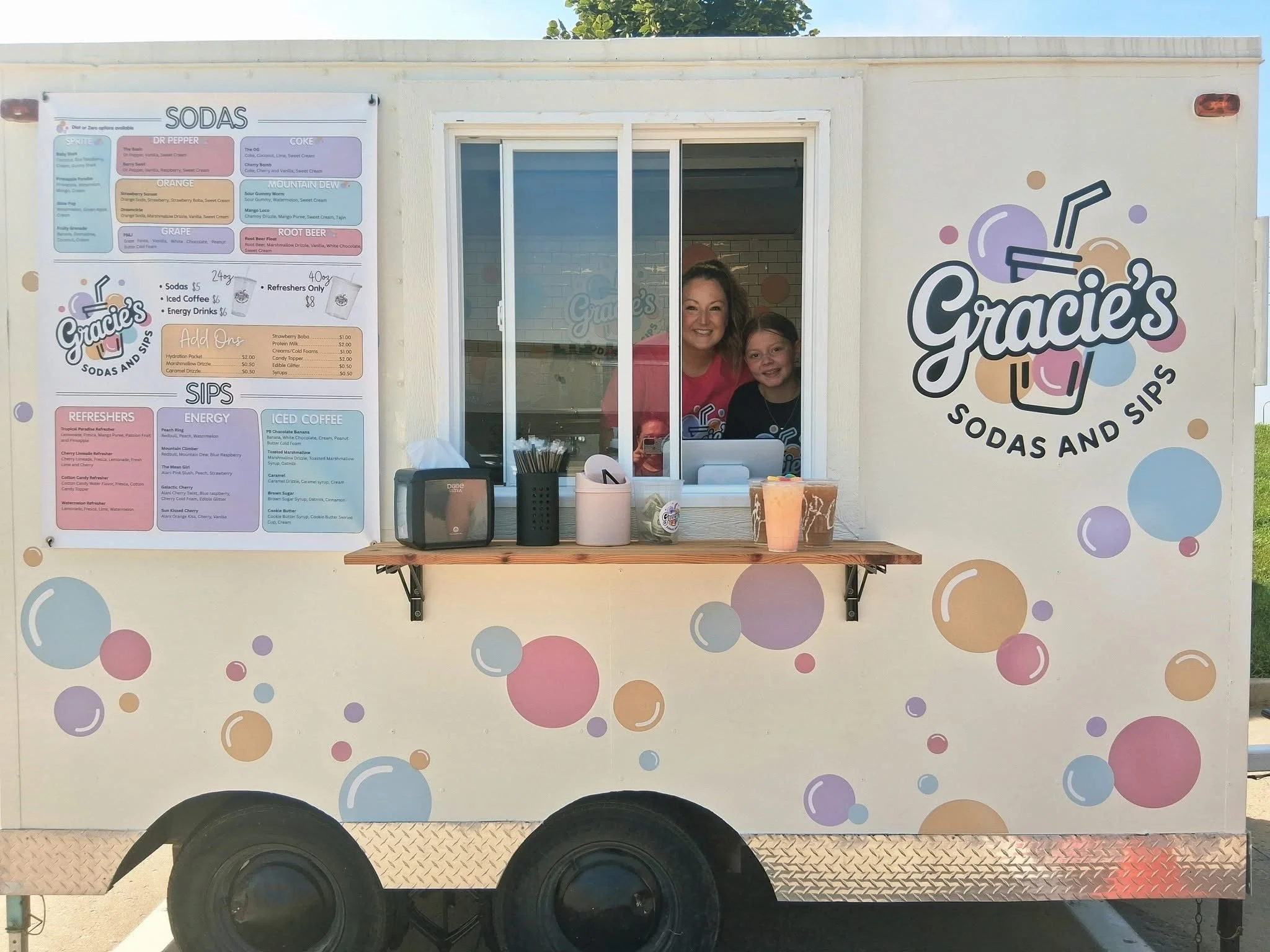 A food truck named Gracie's Sodas and Sips with a colorful bubble pattern on the exterior. There is a window where two women are smiling, selling drinks. The truck has a menu displayed on the left side listing various sodas, sips, and other beverages.