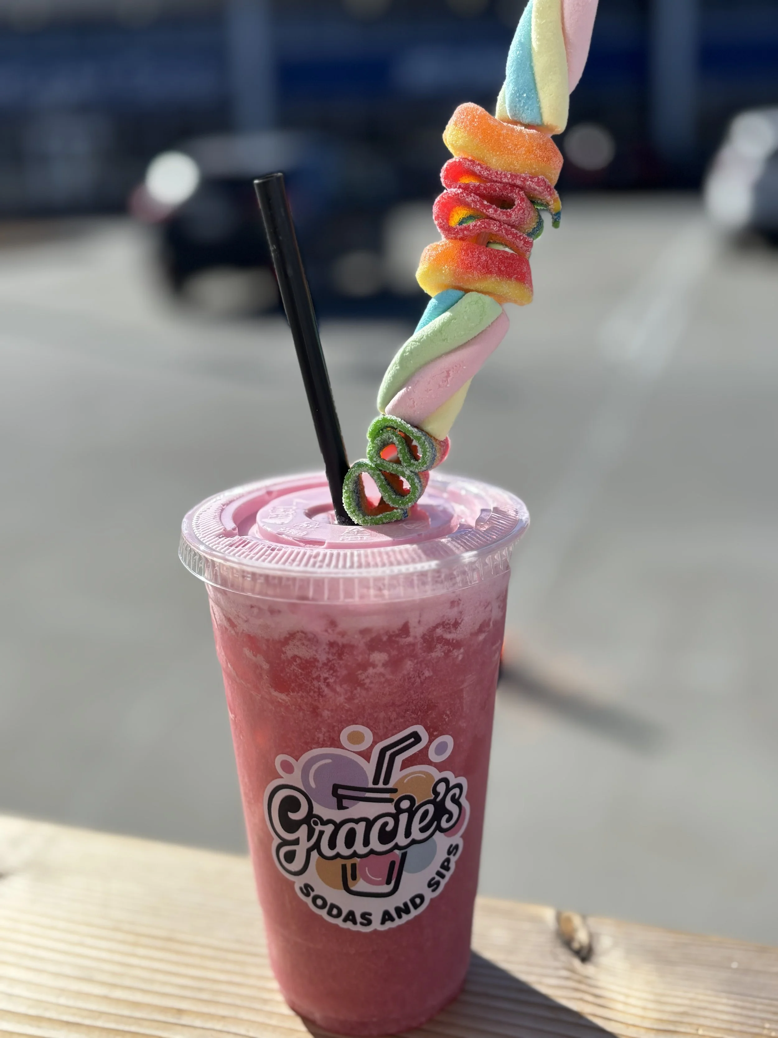 A pink frozen beverage in a clear plastic cup with a black straw and decorated with a multi-colored twisted marshmallow and gummy worms on top.