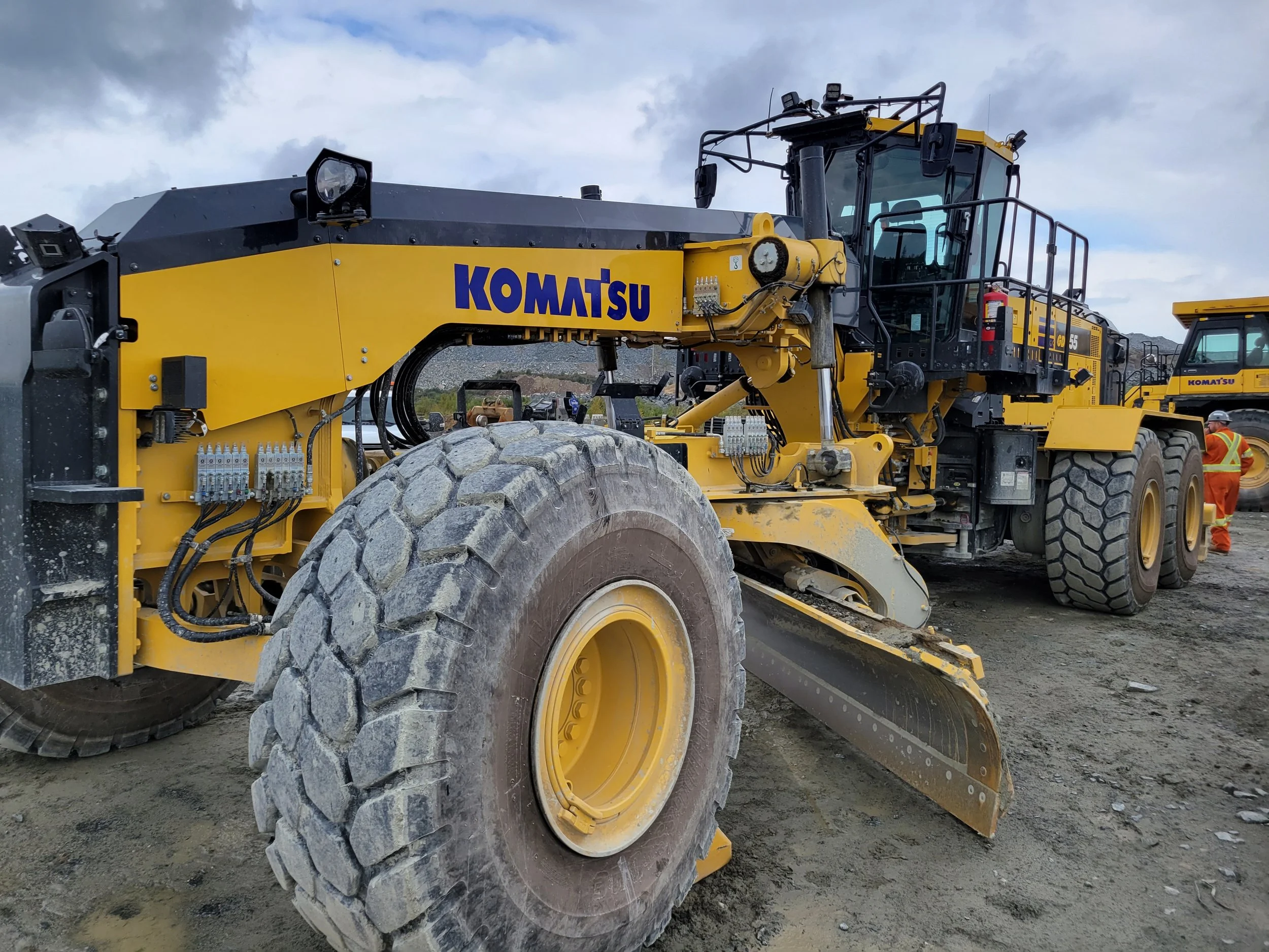 A large yellow Komatsu construction vehicle on a dirt construction site, with another similar vehicle and a worker in safety gear in the background.