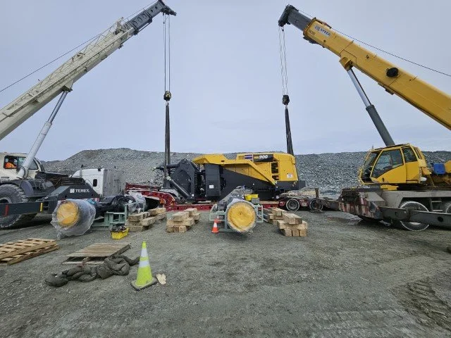 Construction site with two large cranes lifting equipment, a yellow road milling machine, and various construction materials on the ground.
