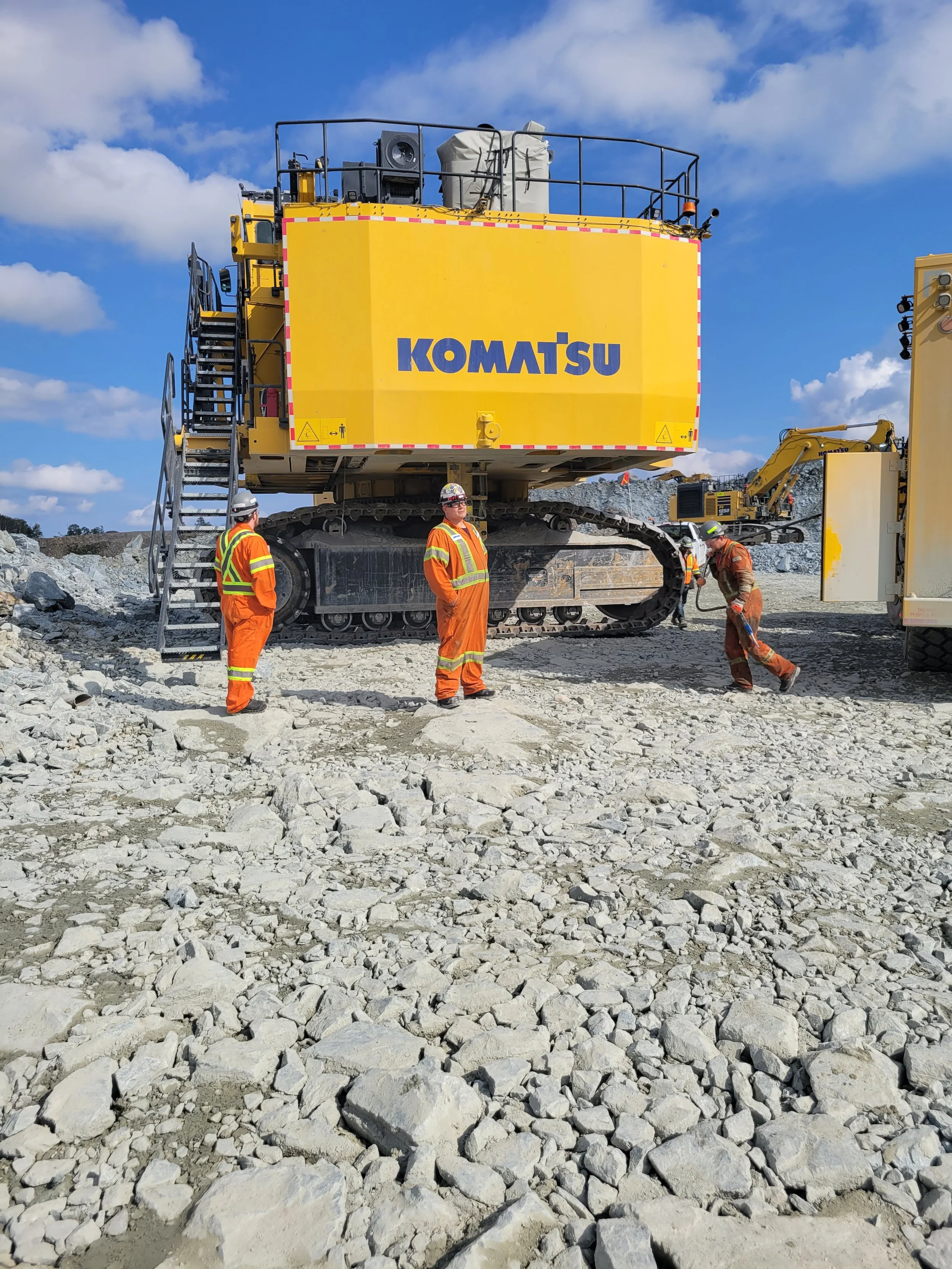 Construction workers in orange safety gear working on rocky terrain near a large yellow Komatsu excavator on a clear day.