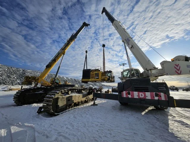 Two large cranes lifting heavy equipment in a snowy outdoor construction or industrial site under a partly cloudy sky.