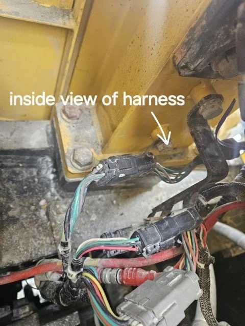 Inside view of a tractor's wiring harness with several colored wires connected to a gray connector, mounted near a yellow metal frame.