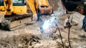 A person welding at a construction site, with sparks flying from the welding process and construction equipment in the background.