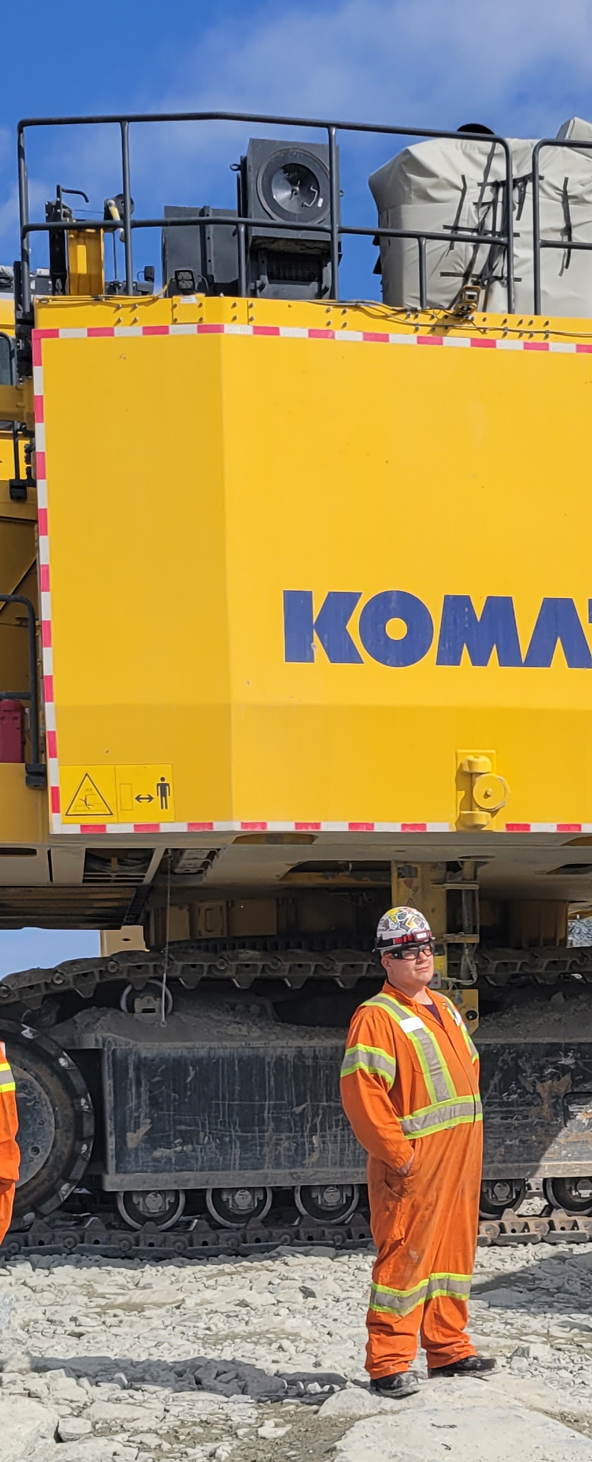 Construction worker in orange safety gear and helmet standing outdoors on rocky ground next to a large yellow Komatsu excavator with a person on top.
