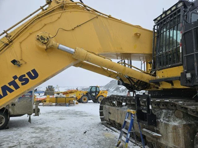 Close-up of a large yellow excavator in a snow-covered construction site, with the arm extended and a black cab, neighboring construction equipment visible in the background.