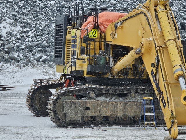 A large yellow excavator with tracked wheels, parked on a rocky terrain, near a rocky hillside.