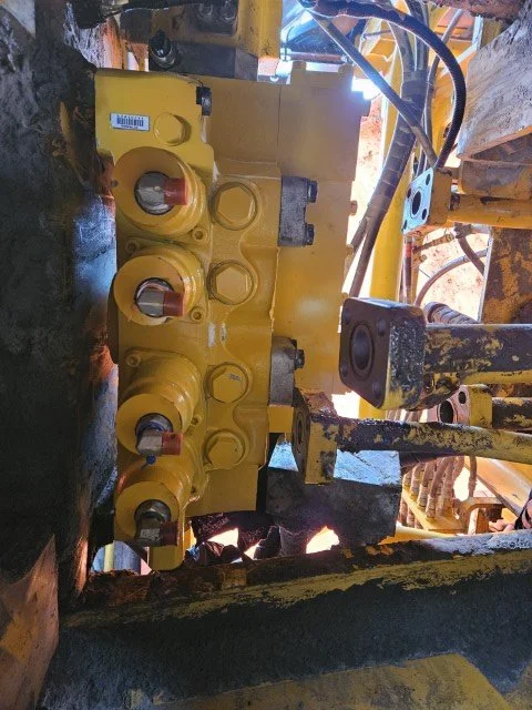 Close-up of a hydraulic control valve assembly on construction equipment, showing multiple yellow valve components and connecting hydraulic hoses.