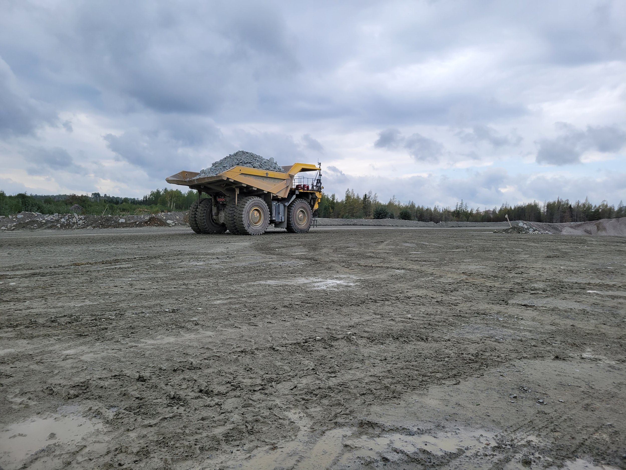 A large yellow dump truck on a construction site, carrying a load of gravel or rocks, with cloudy sky overhead and trees in the background.