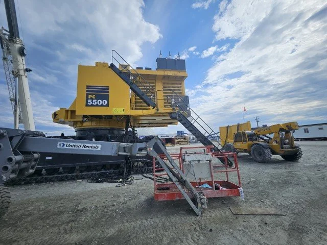Construction site with large yellow PC 5500 rig, a yellow telescopic forklift, a red lift with stairs, and a blue sky with clouds.