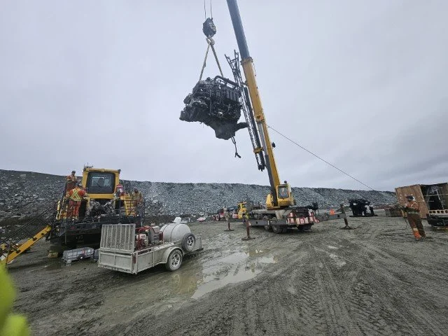 Construction site with a large crane lifting a heavy equipment piece into the air, multiple construction vehicles and workers present.
